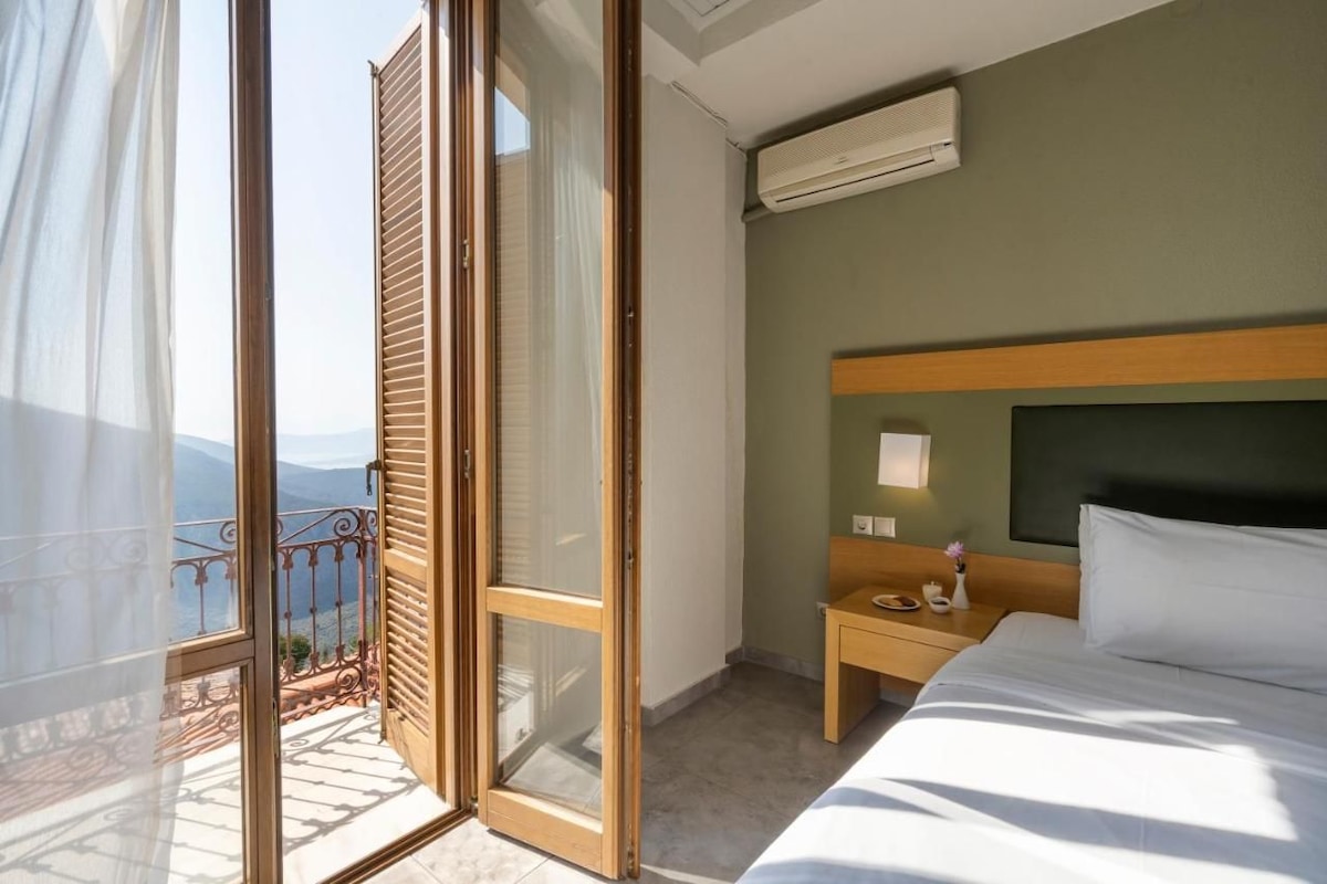 A hotel room with a bed, a nightstand, an air conditioning unit, a window with a view of the mountains, and a balcony with a railing.