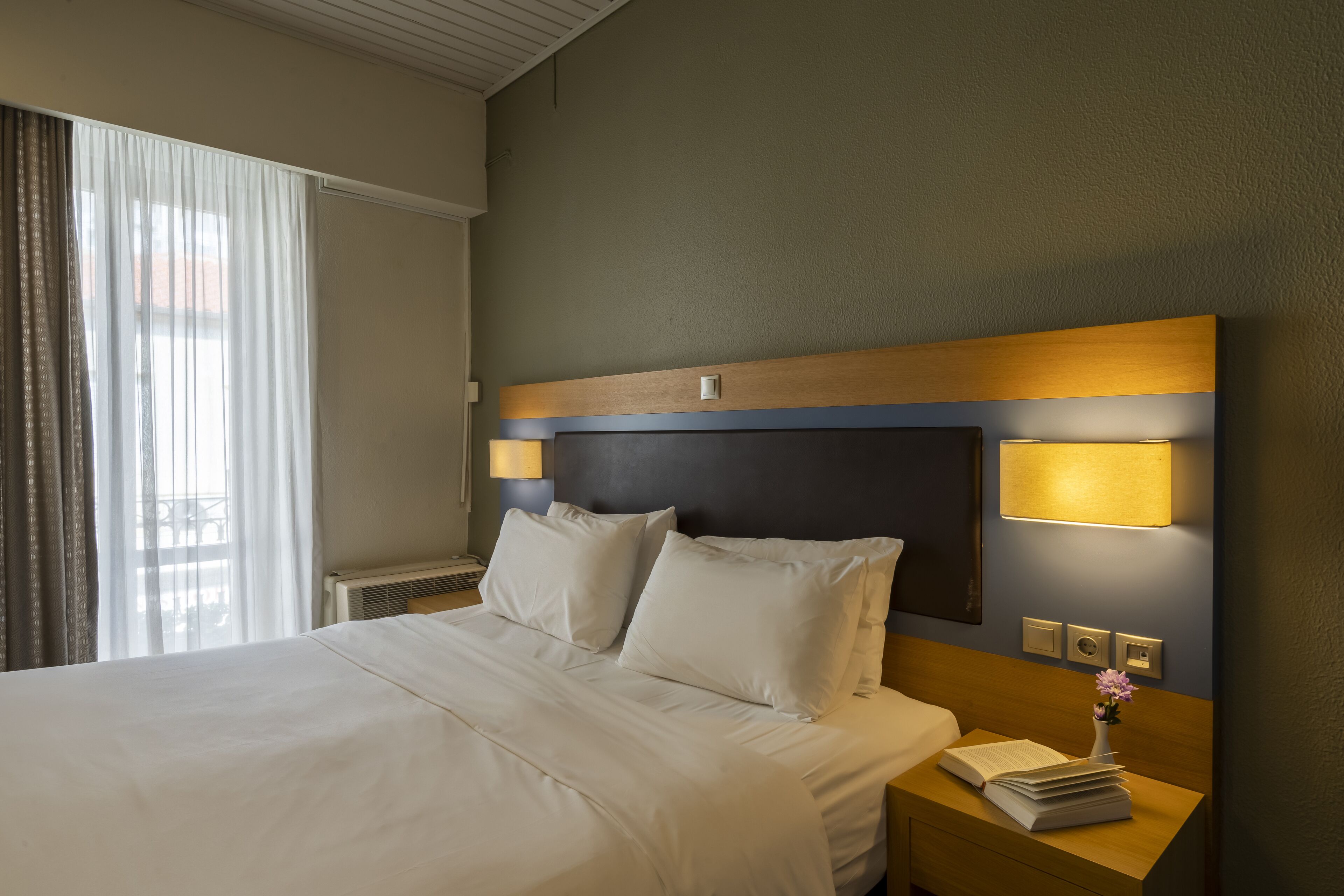 A hotel room with a large bed, two bedside lamps, a nightstand with a book and a vase, and a window with curtains.