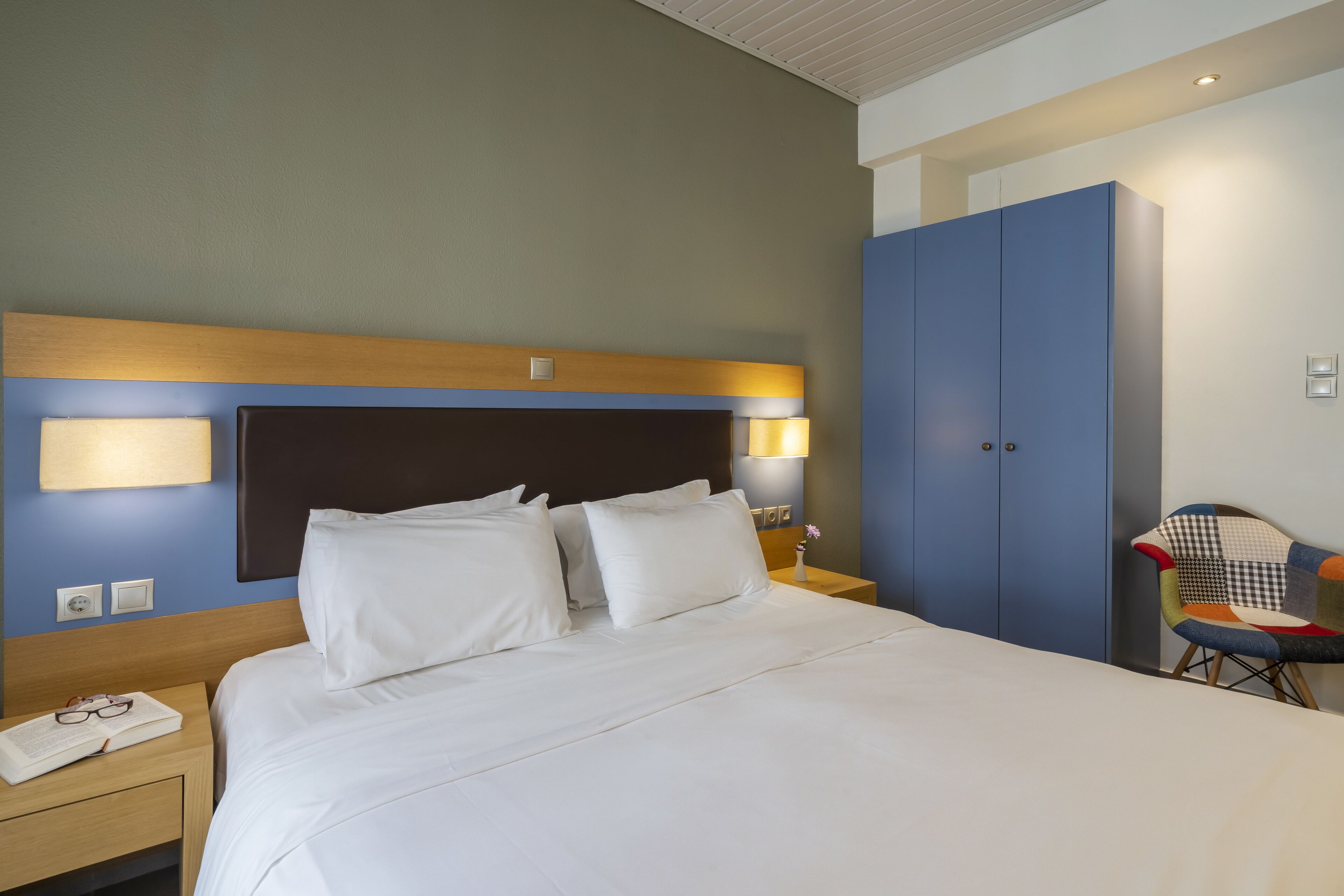 Une chambre d’hôtel avec un grand lit, une table de chevet avec un téléphone et des lunettes, une armoire bleue et une chaise avec un coussin coloré.