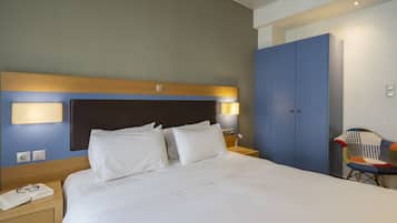 Une chambre d’hôtel avec un grand lit, une table de chevet avec un téléphone et des lunettes, une armoire bleue et une chaise avec un coussin coloré.