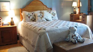 A cozy bedroom with a wooden headboard, a quilted bedspread, a teddy bear, a nightstand with a lamp, and a dresser with a mirror. - Magnetic Hill Winery (Moncton)