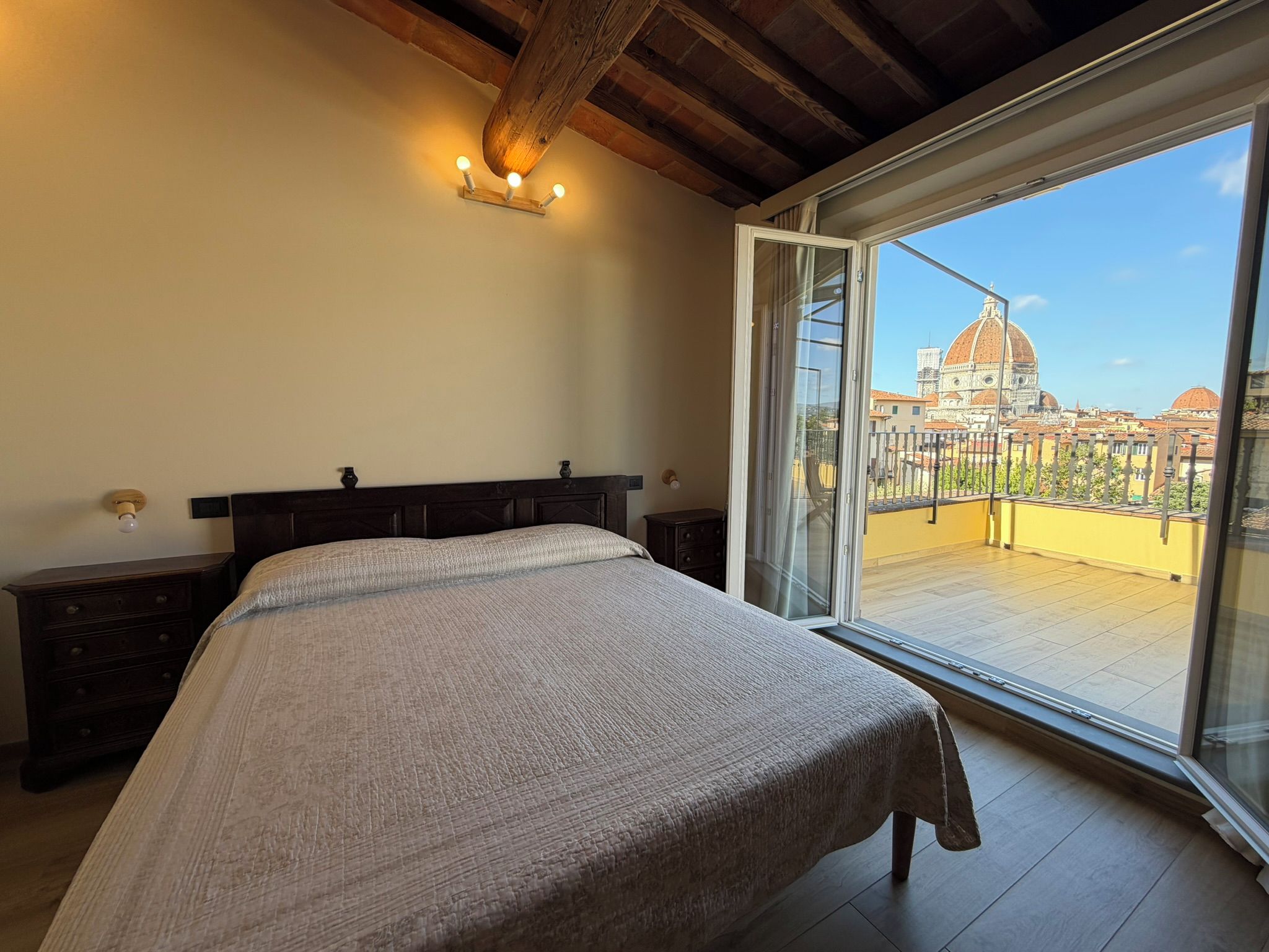 Une chambre avec un grand lit, une tête de lit en bois, une table de chevet, une lampe de chevet et une vue sur le paysage urbain, visible à travers une porte-fenêtre coulissante.