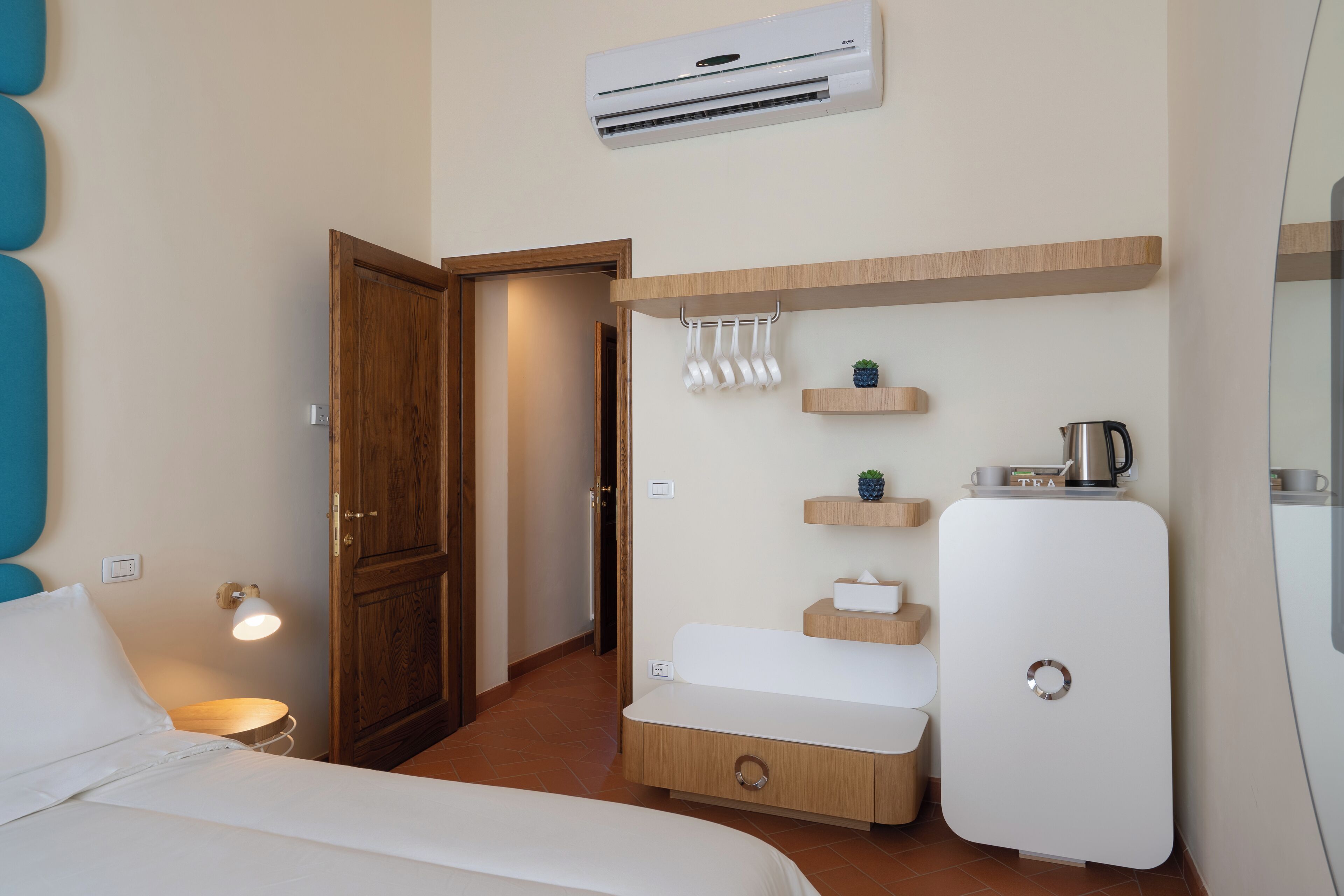 A compact hotel room with a bed, a small bedside table, a built-in wardrobe, a mini-fridge, and a wall-mounted air conditioner.