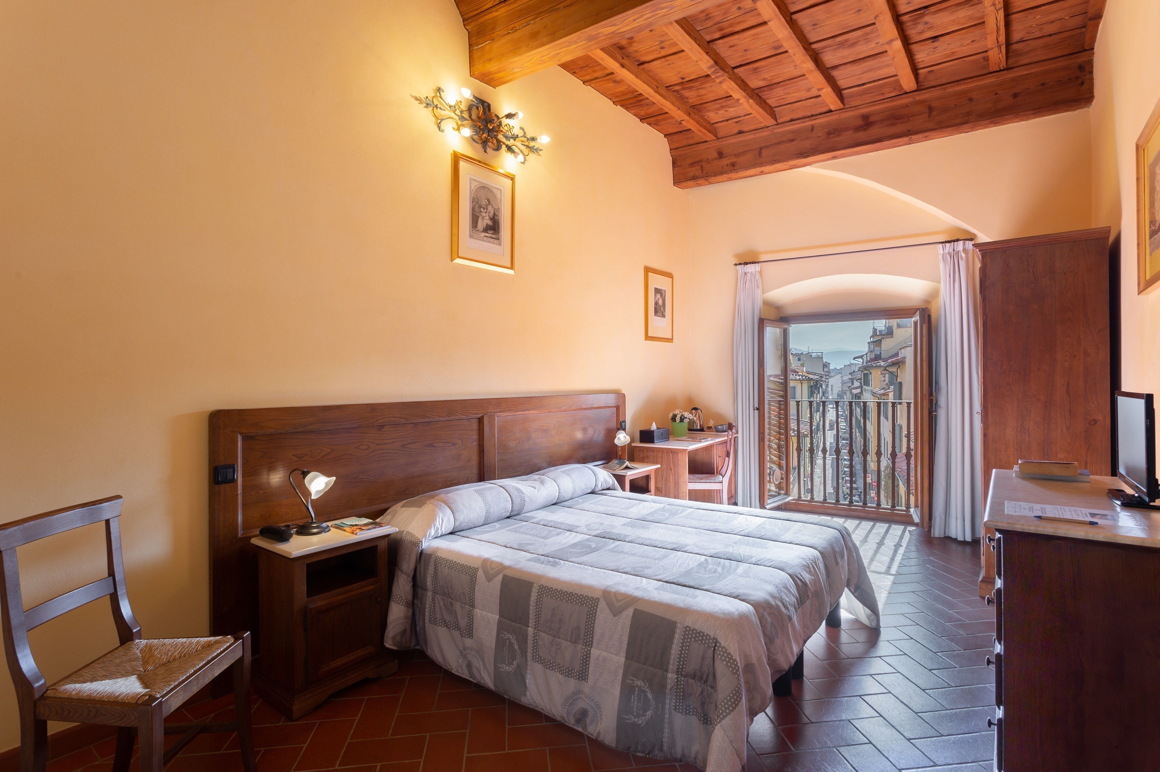 A bedroom with a wooden headboard, a bed with a patterned bedspread, a nightstand with a lamp, a wooden desk, and a view of a cityscape through a balcony door.