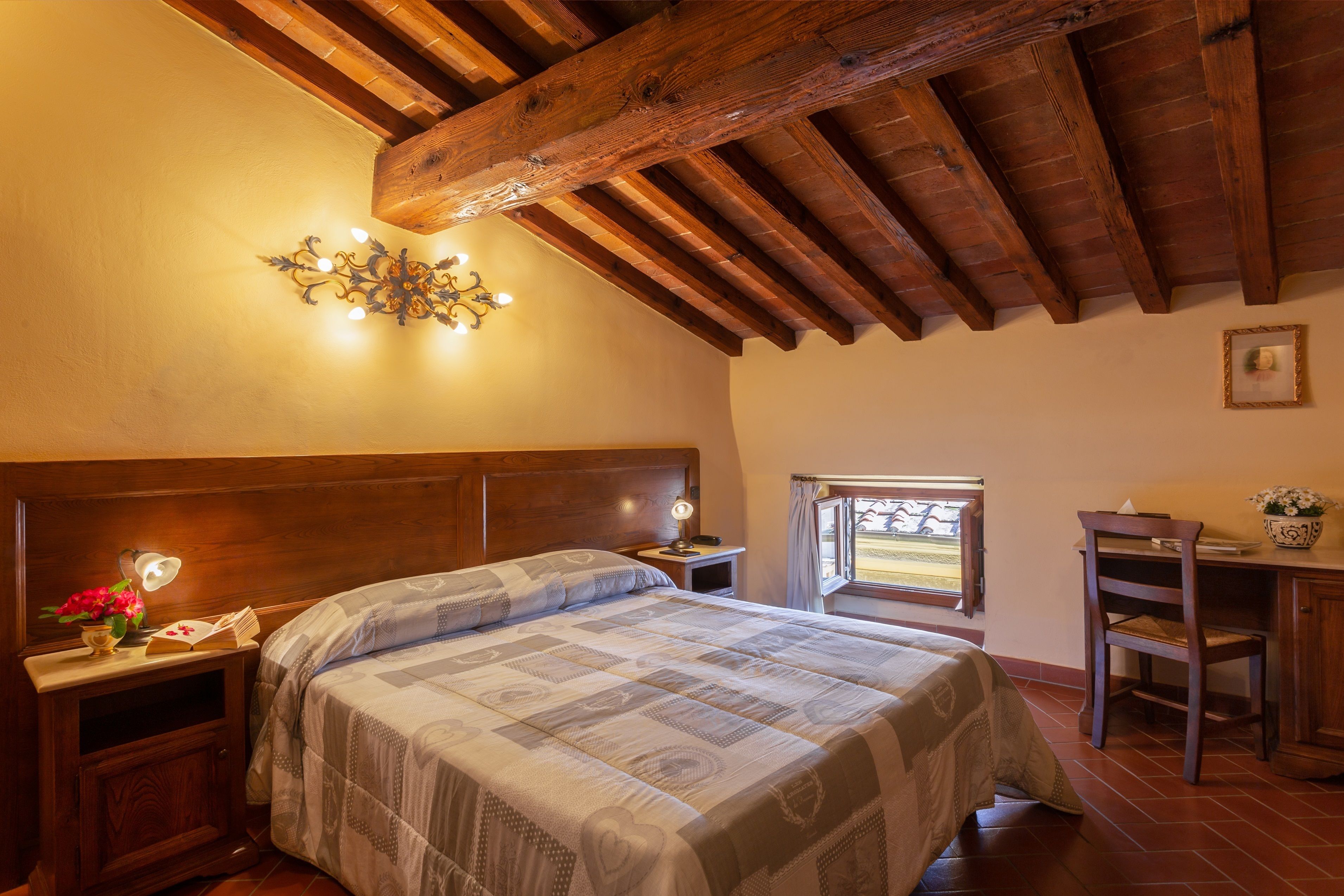 A bedroom with a wooden ceiling, a bed with a patterned bedspread, a nightstand with a lamp, and a desk with a chair.