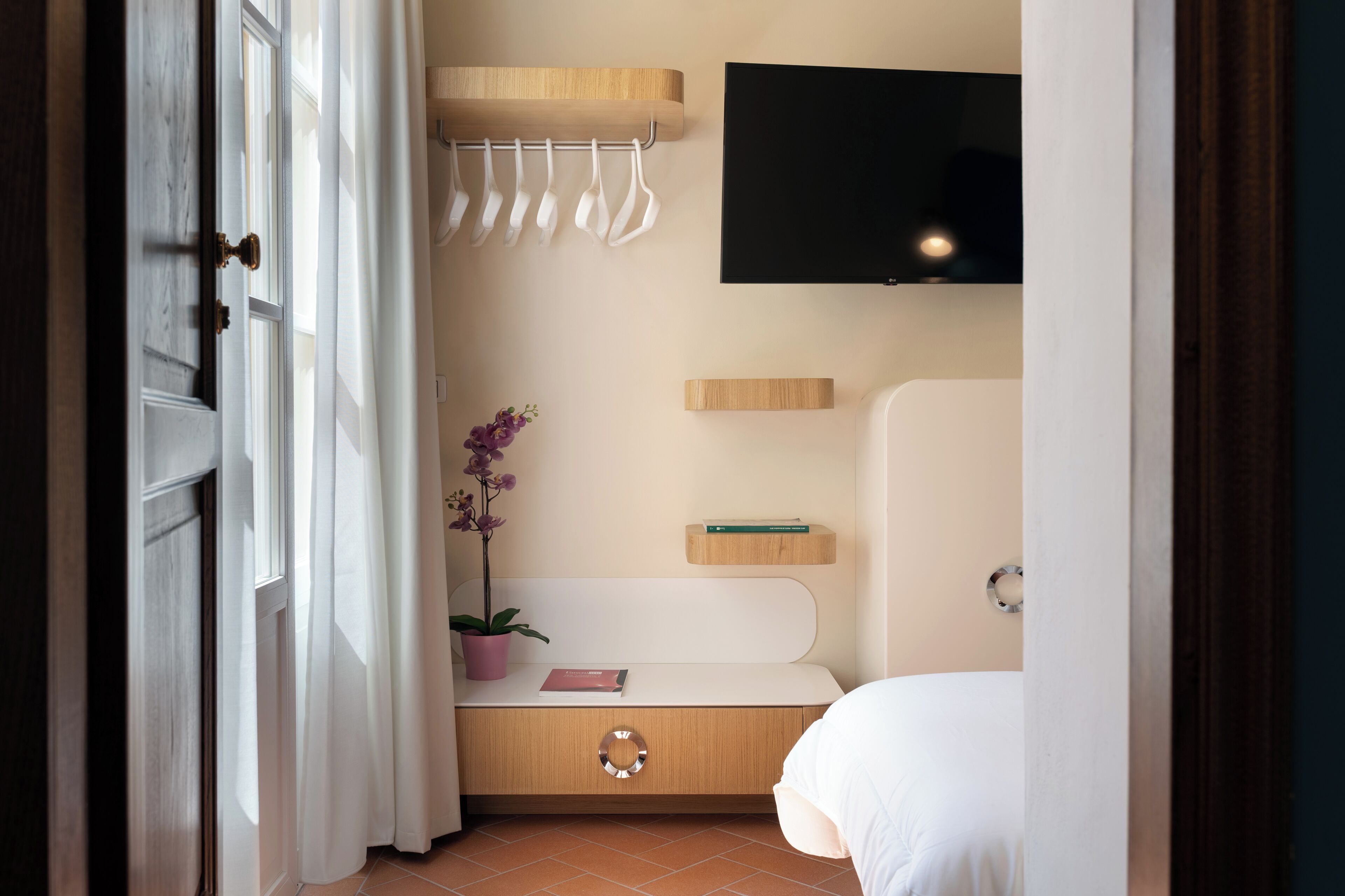 Een hotelkamer met een bed, een aan de muur bevestigde televisie, een klein plankje met boeken, een kamerplant en een handdoekhouder.