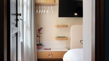 Een hotelkamer met een bed, een aan de muur bevestigde televisie, een klein plankje met boeken, een kamerplant en een handdoekhouder.