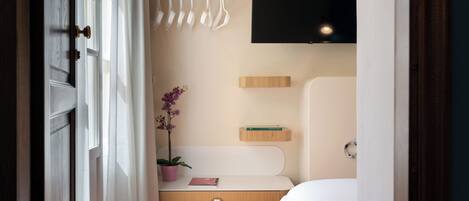 A hotel room with a bed, a TV mounted on the wall, a small shelf with books, a potted plant, and a towel rack.
