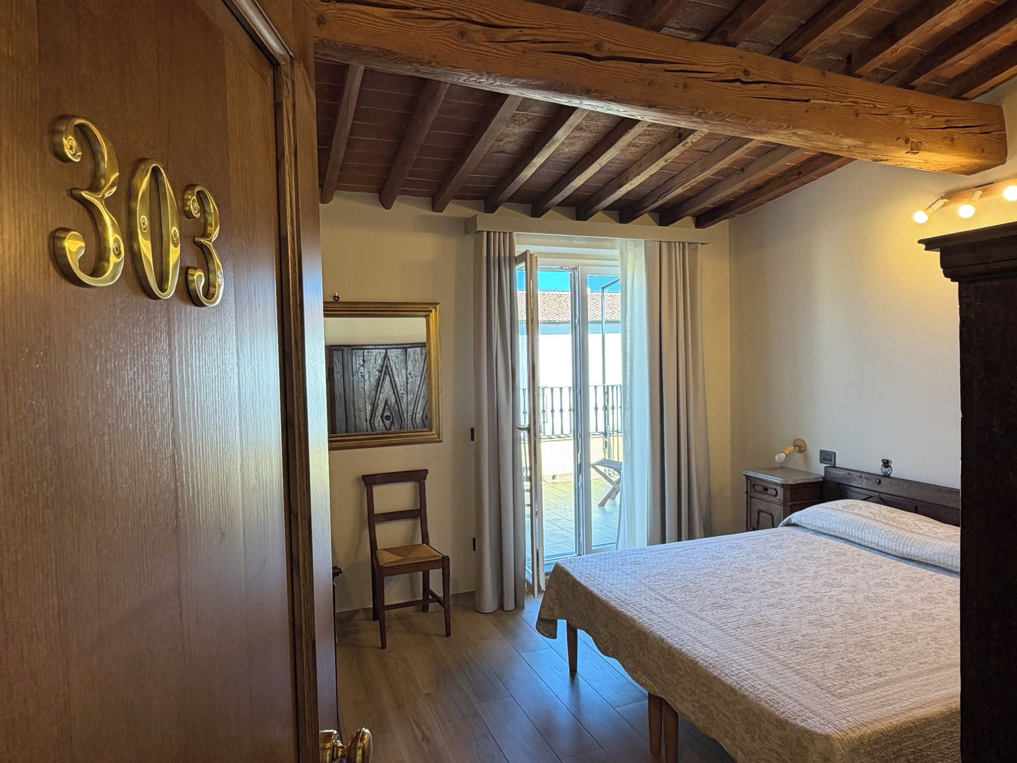 Une pièce avec un lit, une chaise, une porte en bois portant des numéros, et une vue par la fenêtre.