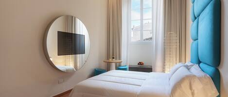 A modern hotel room with a large bed, a round mirror, a small table with a drink, and a view of the city through the window.