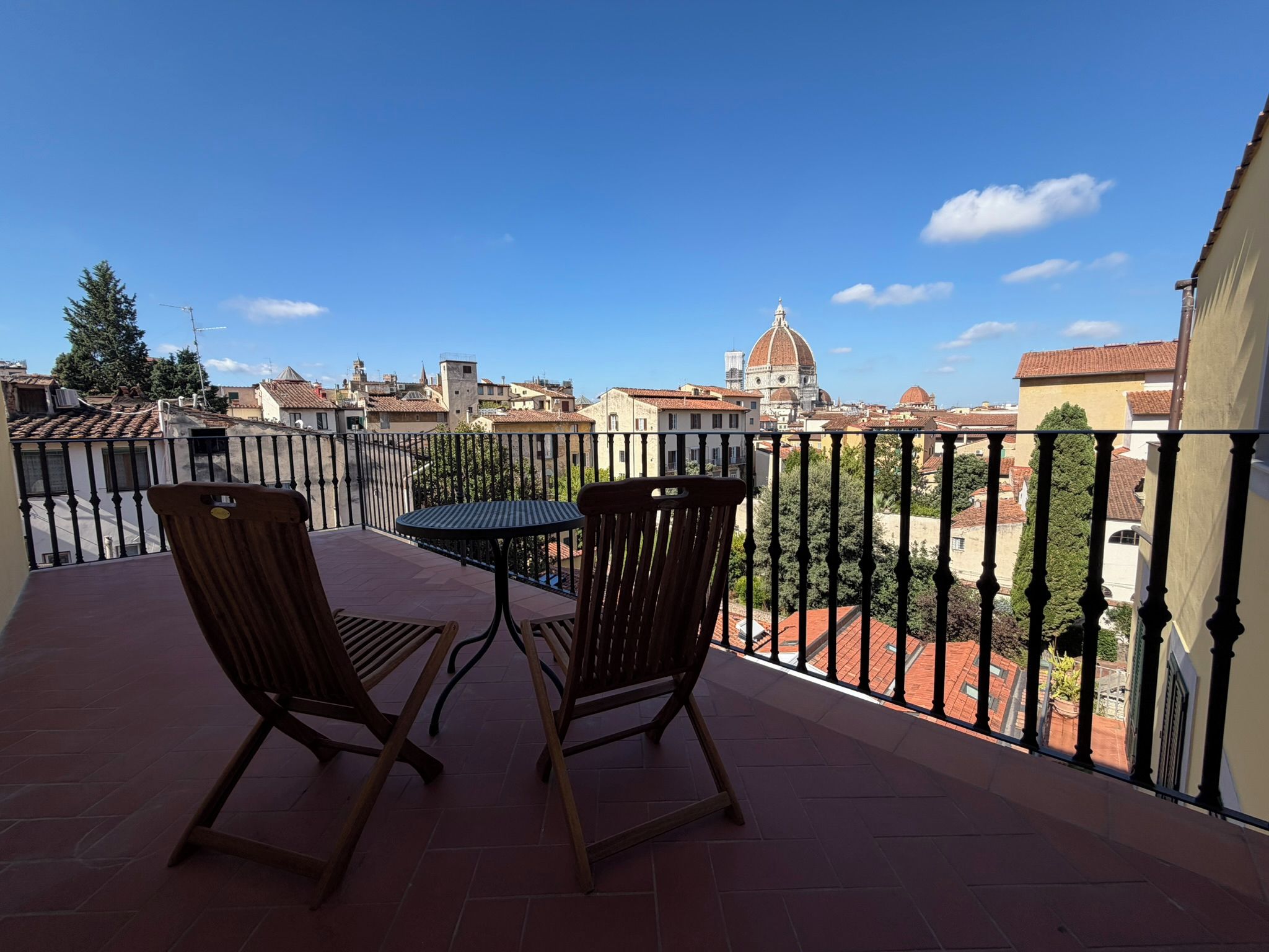 A balcony with two wooden chairs and a table, overlooking a cityscape with historic buildings and a prominent dome.
