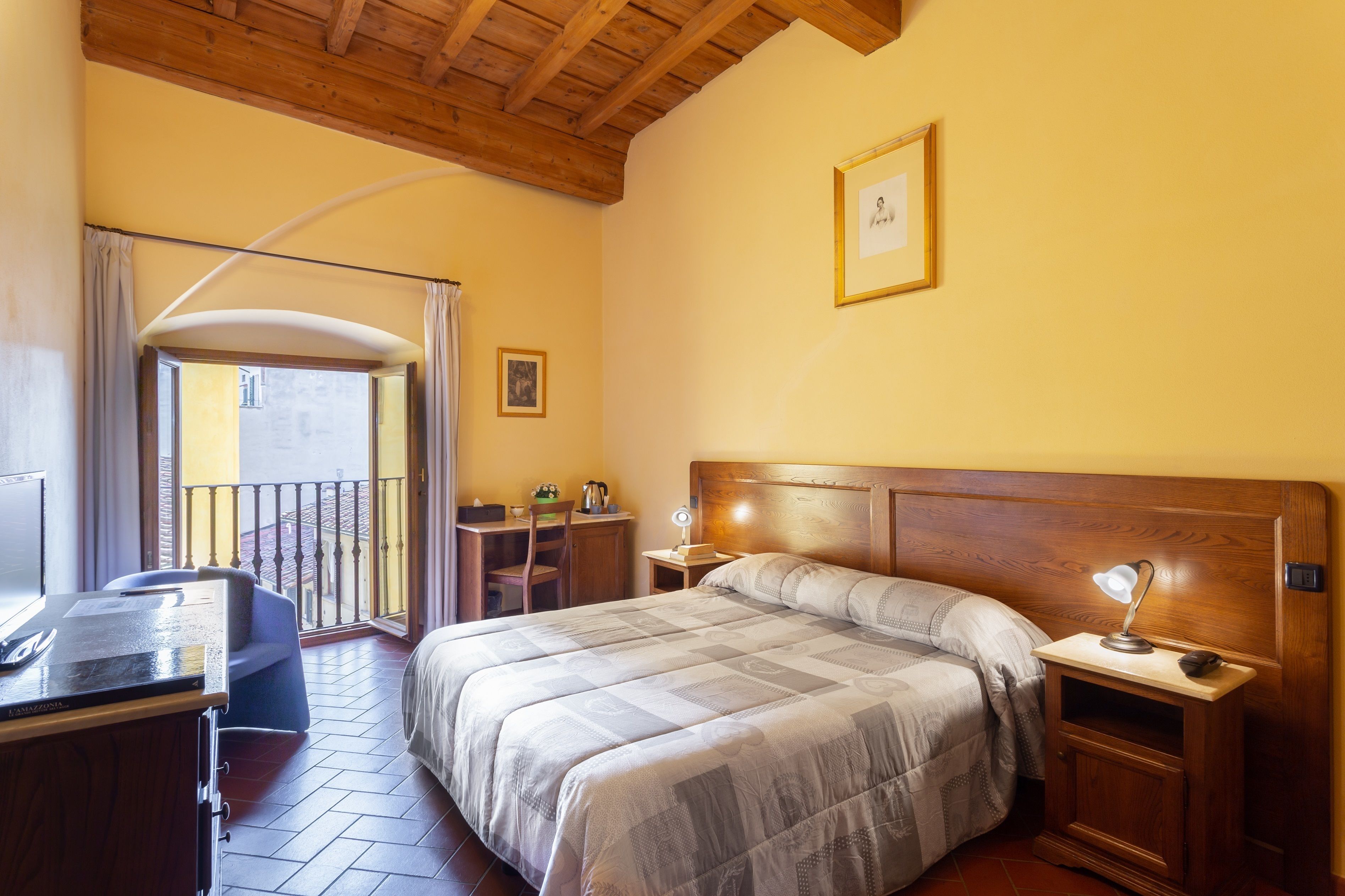 Un dormitorio con cabecera de madera, una cama con colcha, un escritorio con silla, una mesita de noche con lámpara y un balcón con barandilla.