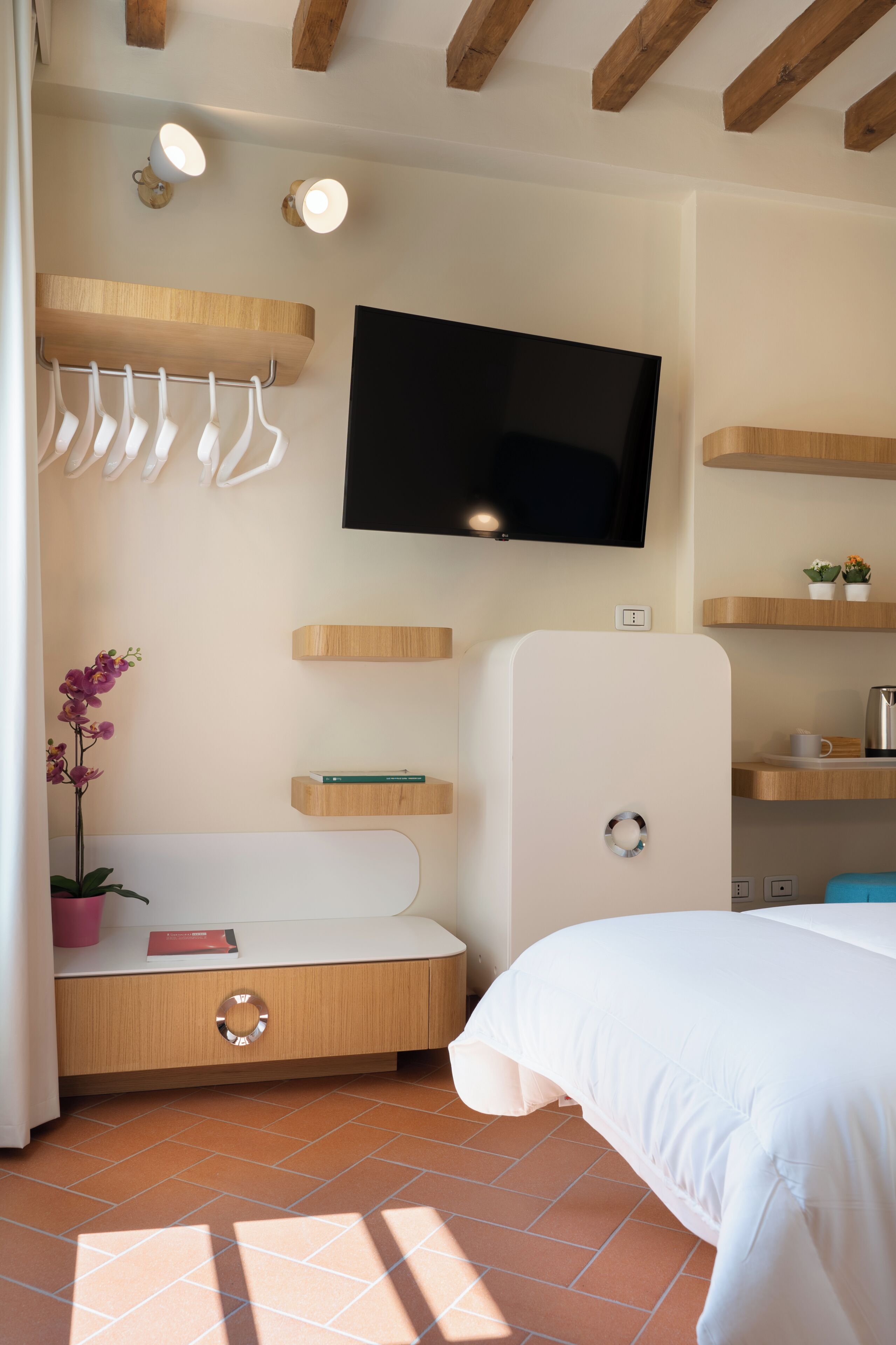 A bedroom with a bed, a TV mounted on the wall, a wooden bench with a plant, and a tiled floor.