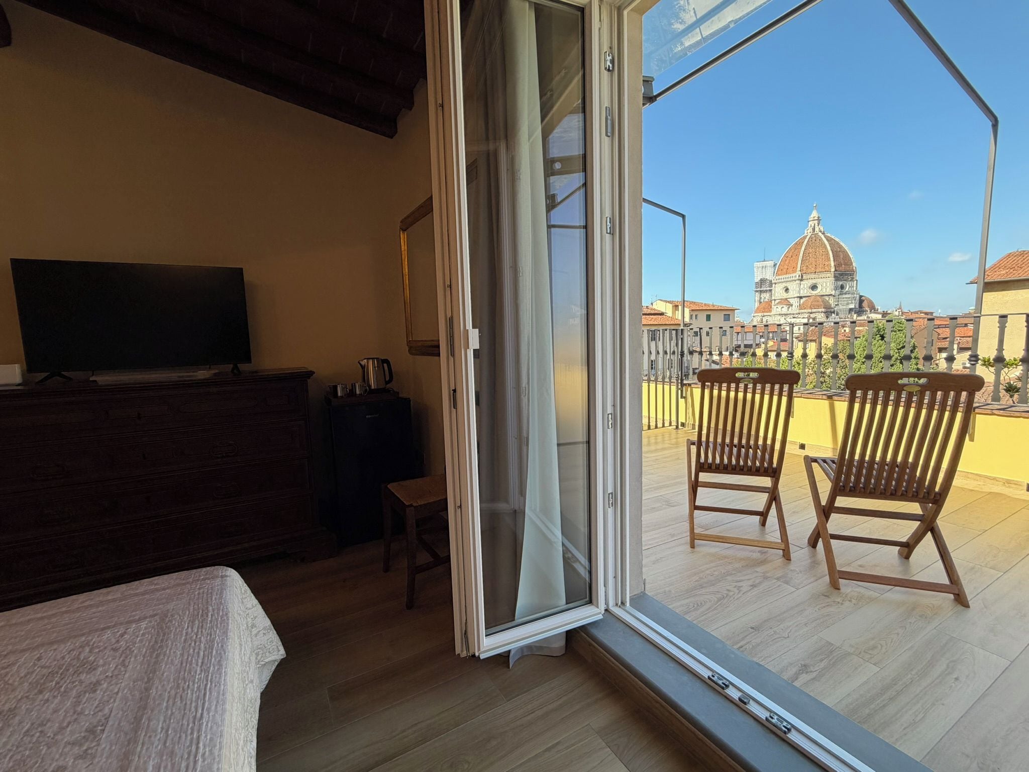 Una habitación con cama, televisión y balcón con dos sillas de madera y vista a un edificio histórico.