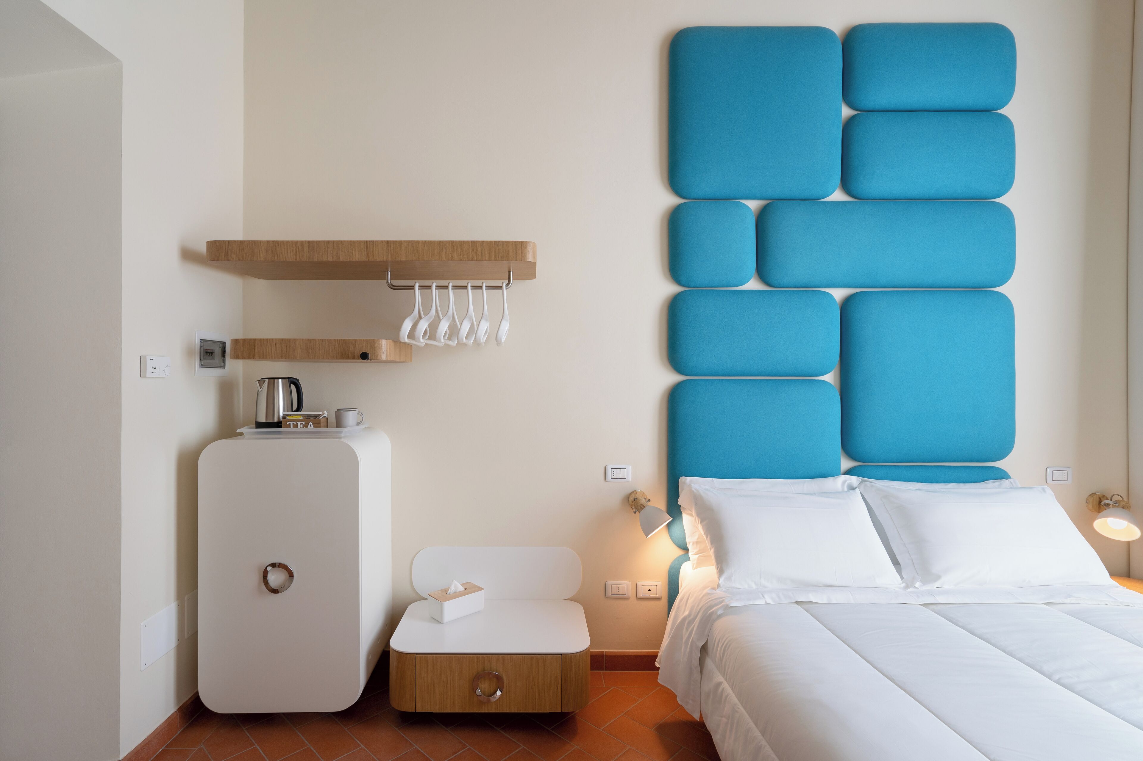 A modern hotel room with a bed, a bedside table with a lamp, a small cabinet, and a wall-mounted shelf with towels.
