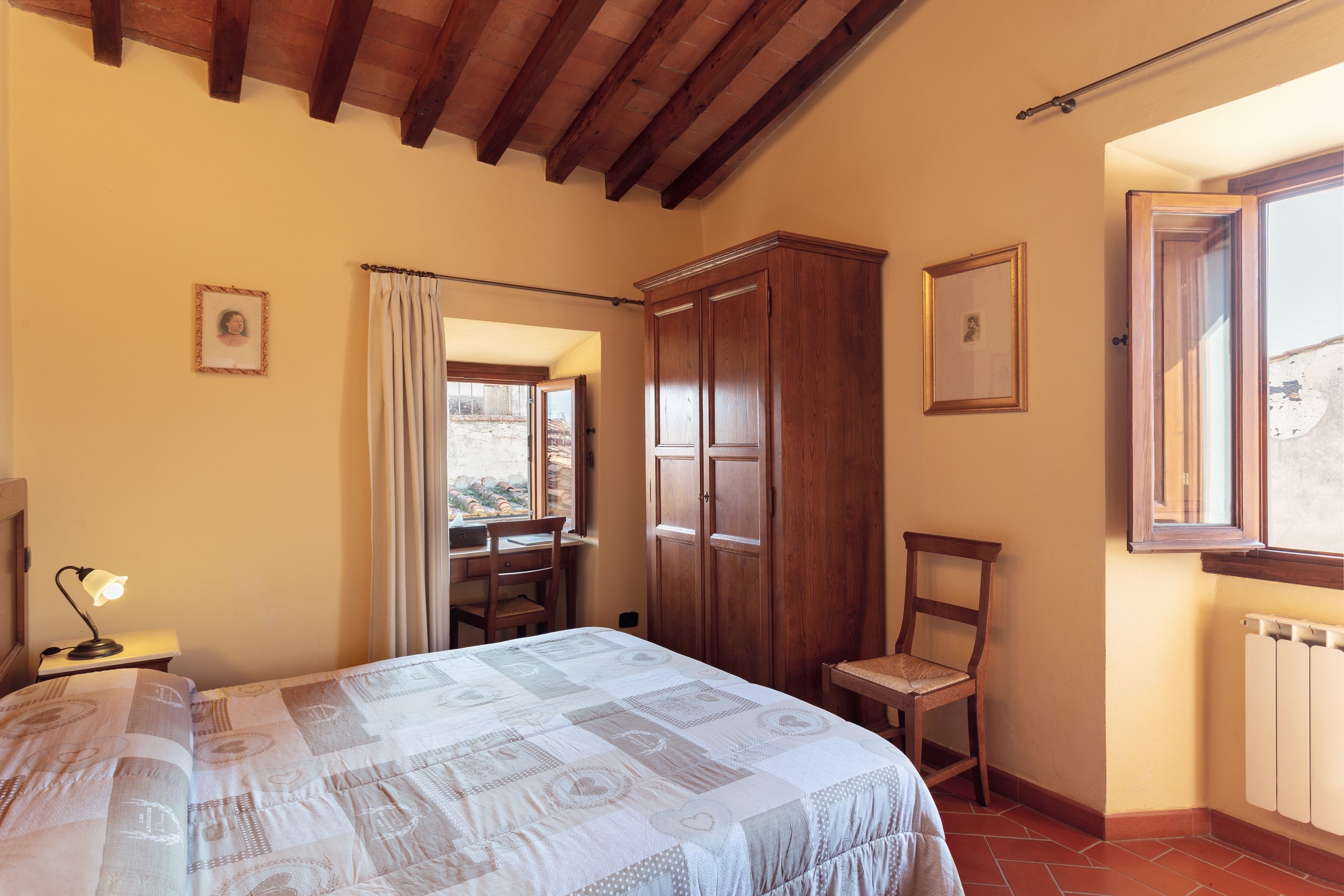 Un dormitorio con cama, armario de madera, una silla, escritorio con lámpara y ventana con cortinas.