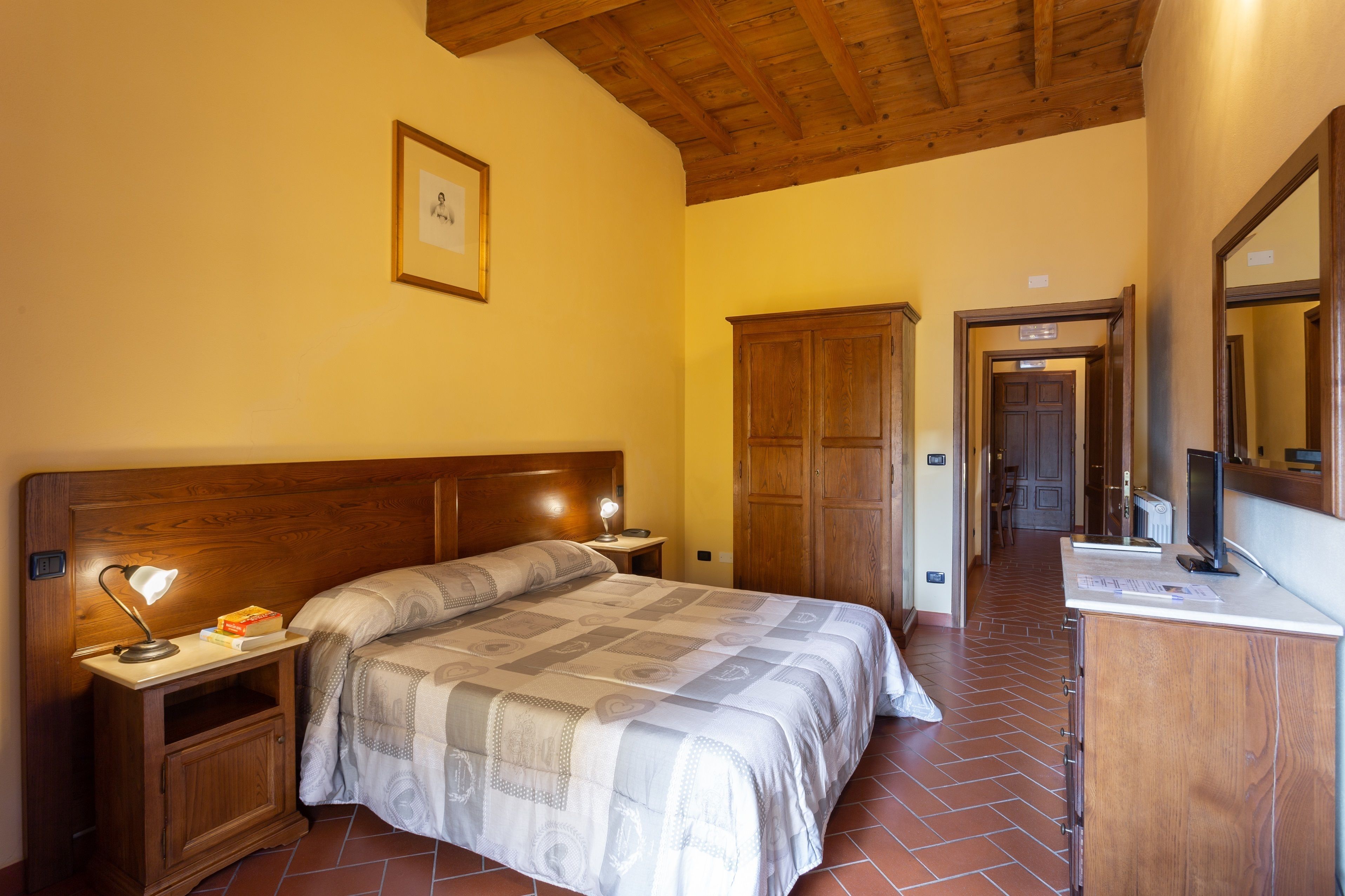 A bedroom with a wooden headboard, a bedside lamp, a bed with a patterned bedspread, a nightstand, a wardrobe, and a mirror.