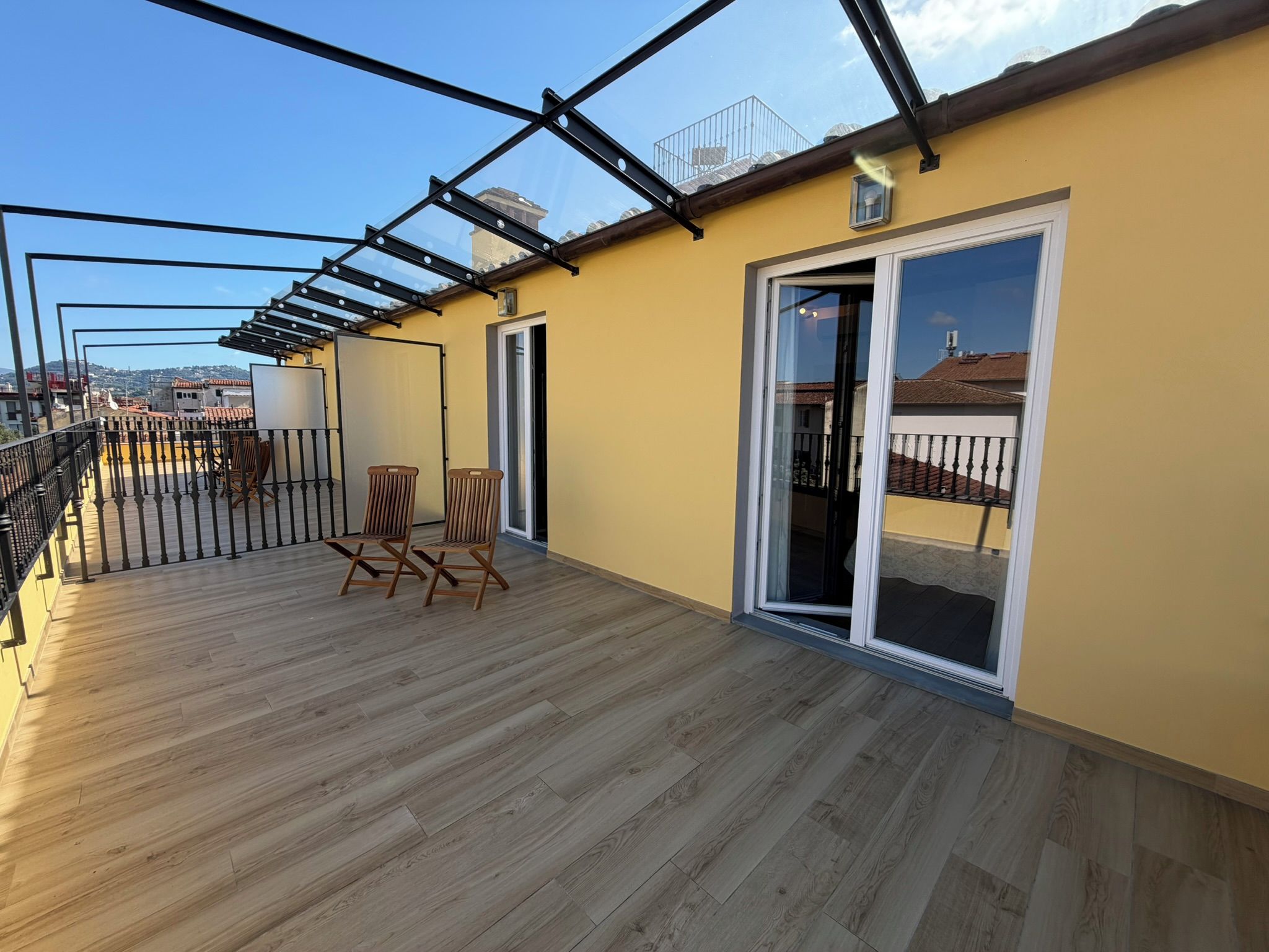 Un balcon doté d’un plancher en bois, de deux chaises en bois, d’une porte vitrée et d’une rambarde en métal.