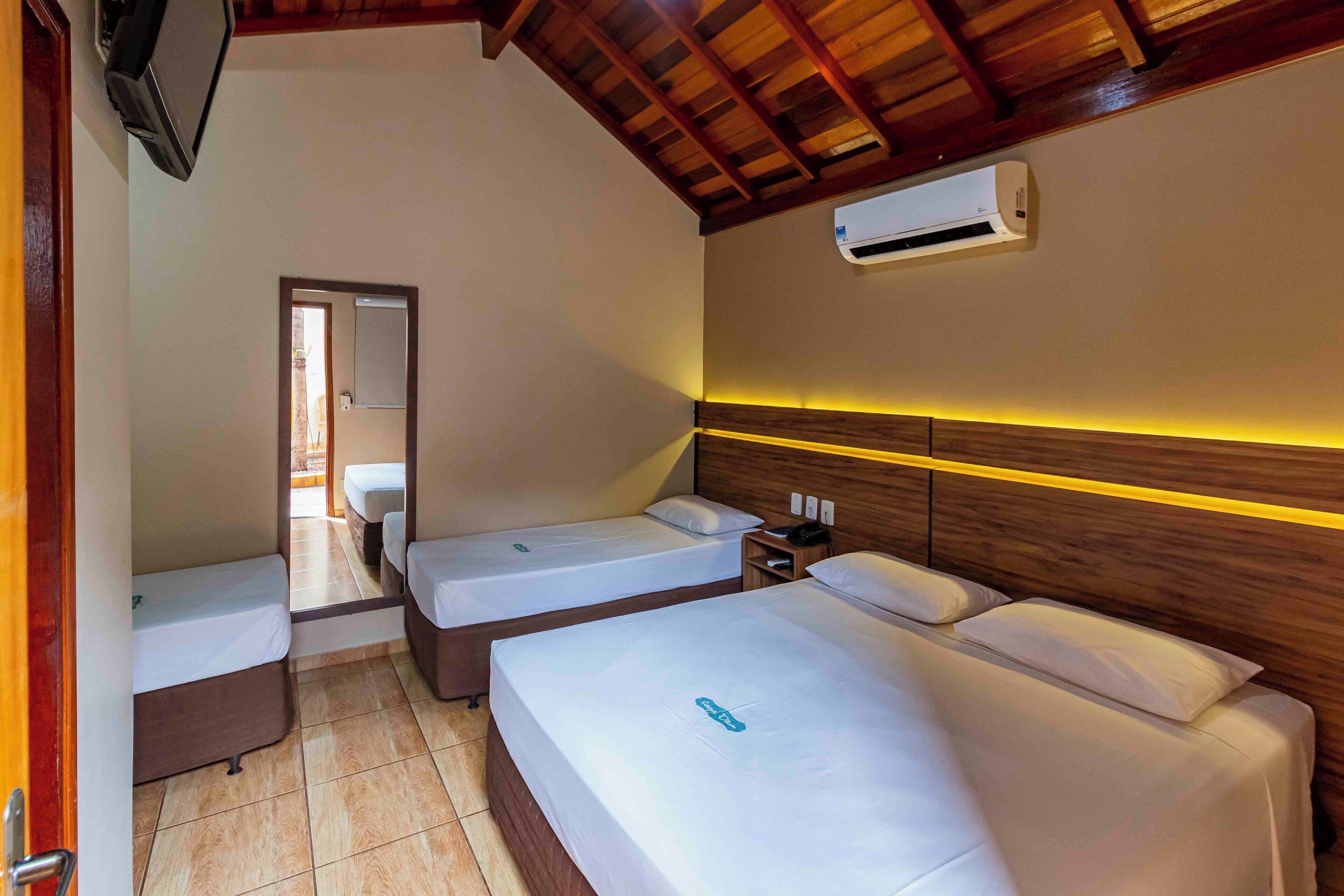 Habitación de hotel con dos camas individuales, cabecera de madera, unidad de aire acondicionado, mesita de noche con lámpara y espejo en la pared.