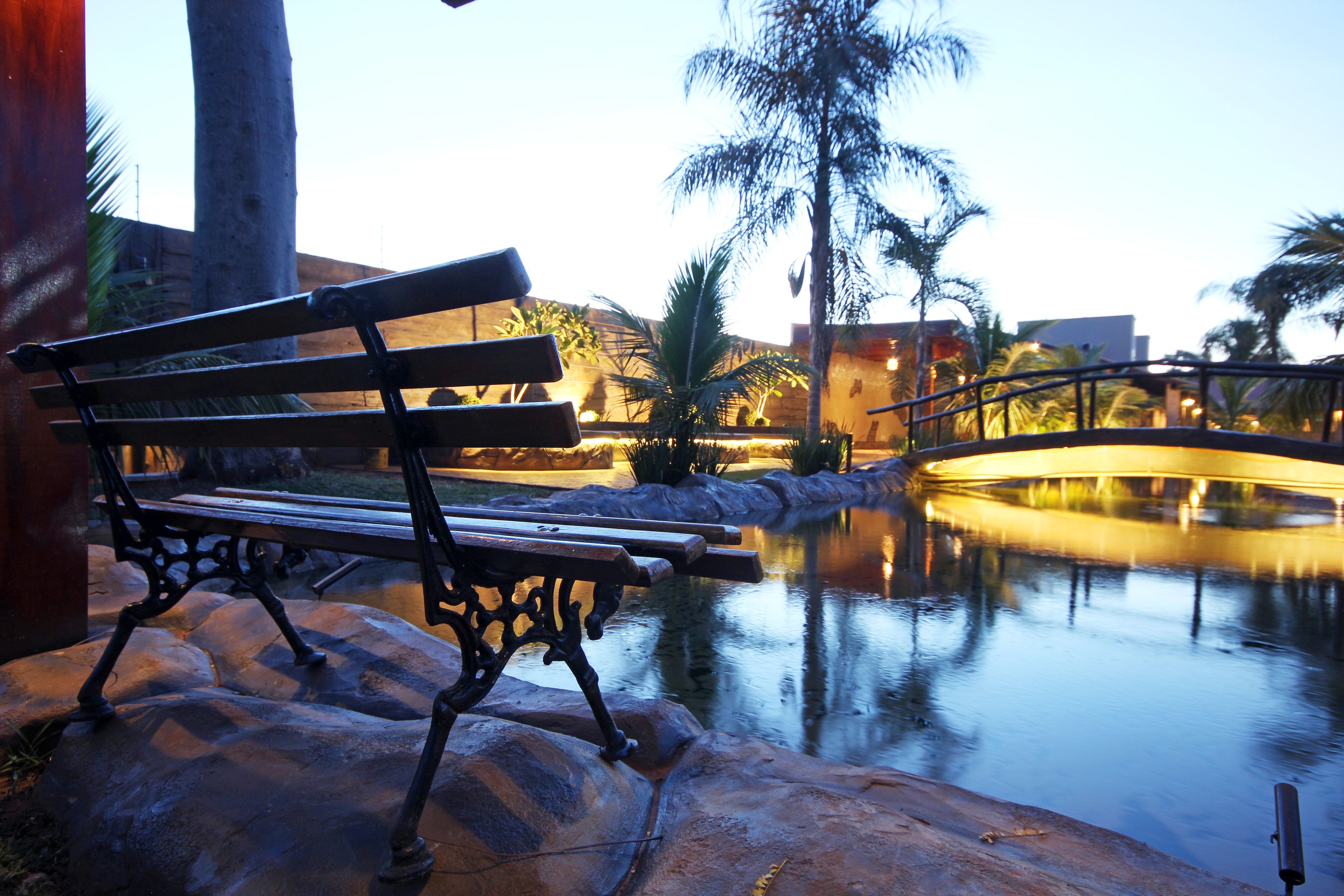 A bench by a calm water feature with a bridge and palm trees in the background.
