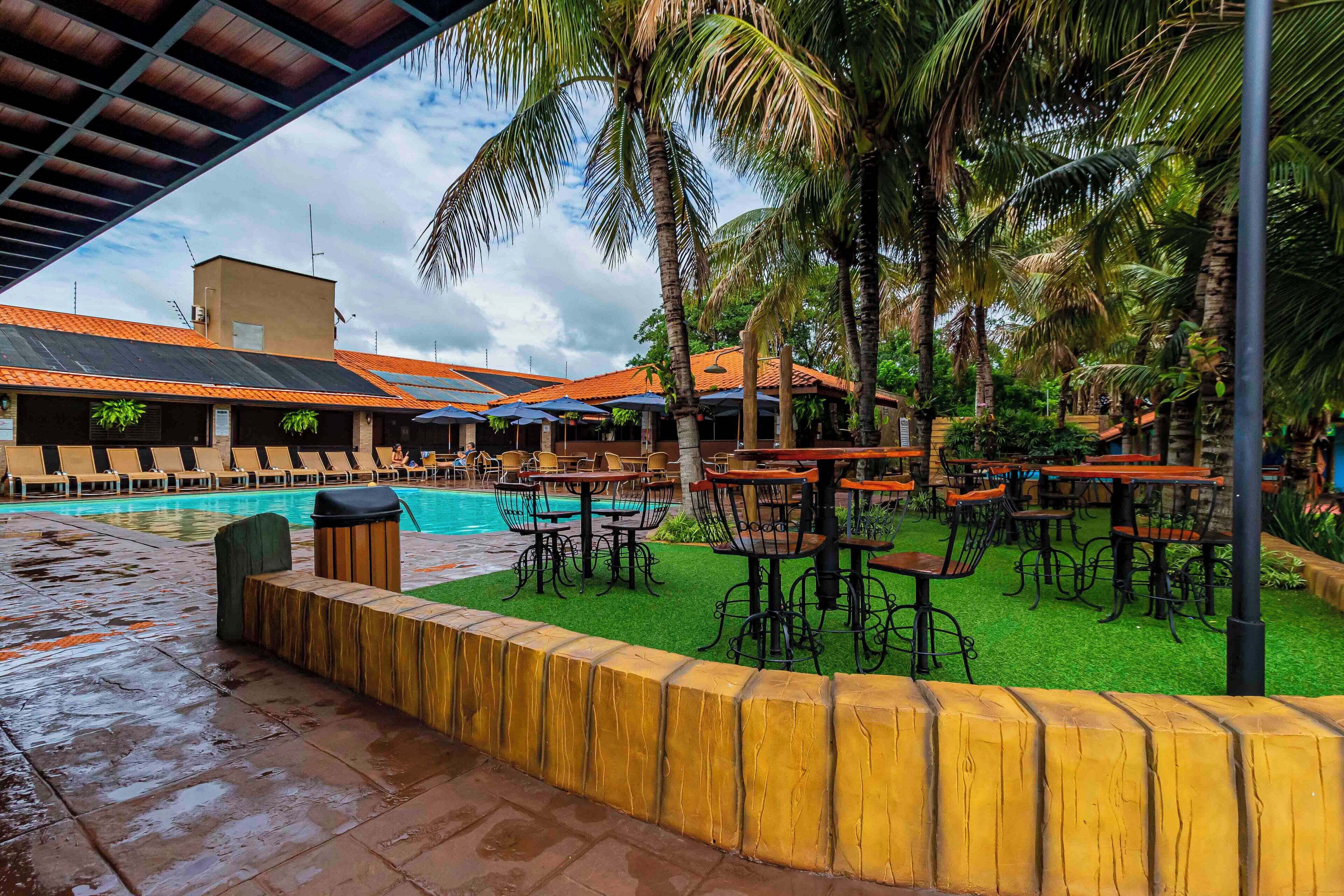 A poolside area with lounge chairs, tables, and umbrellas, surrounded by palm trees and a covered seating area.