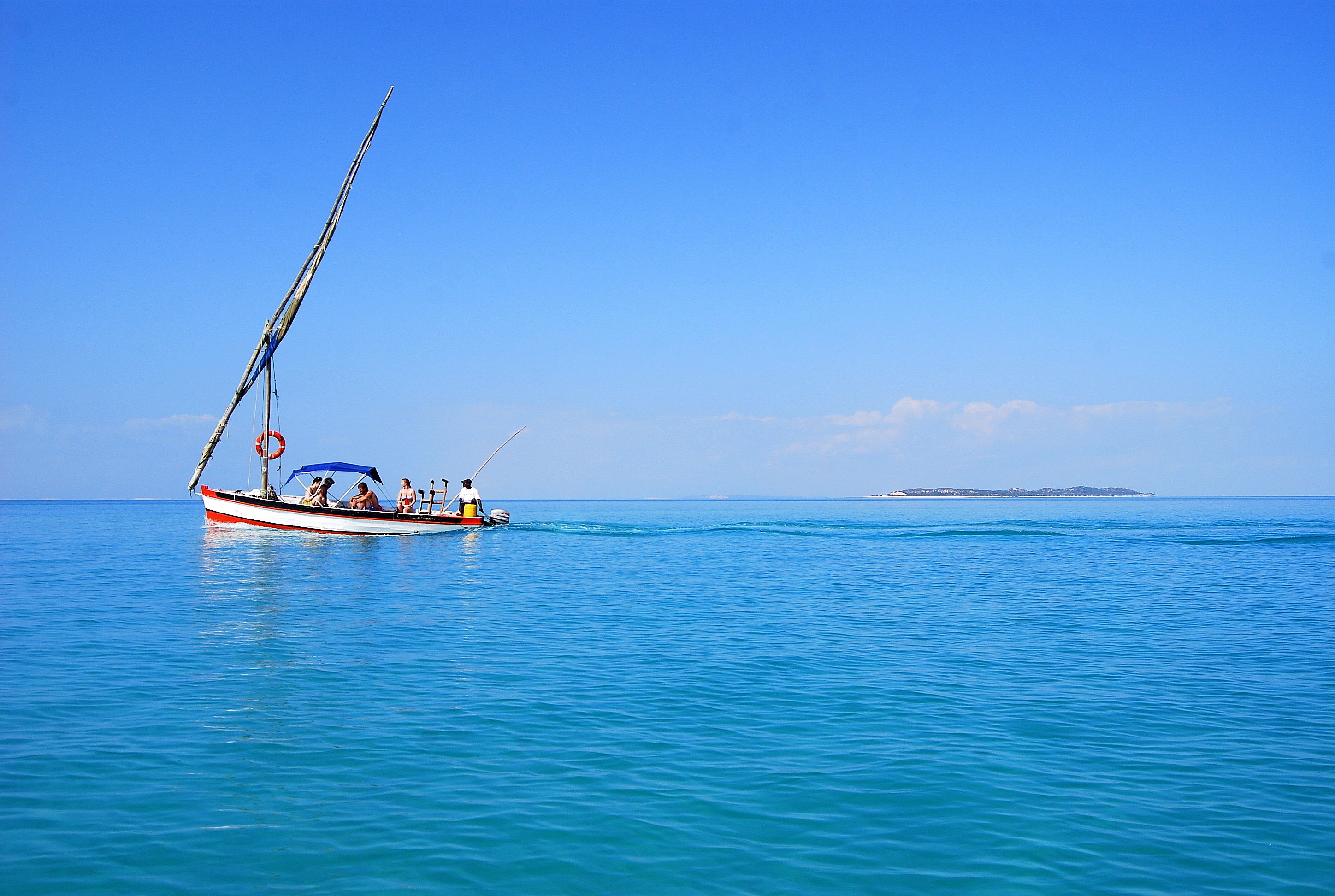 A boat with a sail on calm blue water, a small island in the distance, and a clear sky.