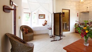Ein Zimmer mit einem Bett, einem Korbstuhl, einem Holztisch und einem durch eine Tür einsehbaren Badezimmer.