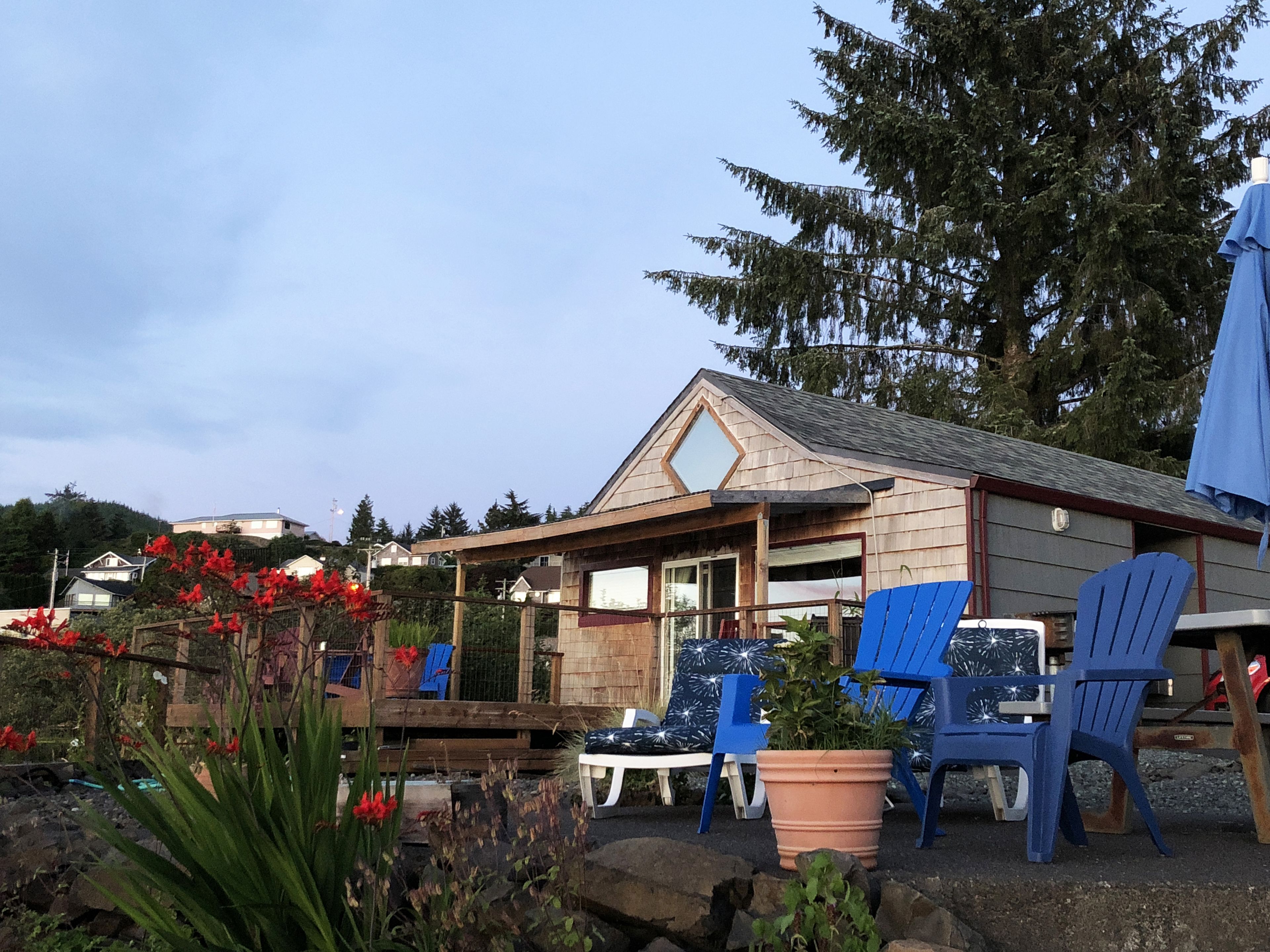 Ein Holzhaus mit geneigtem Dach, einer Sitzgelegenheit im Freien mit blauen Stühlen und einem Garten mit roten Blumen.