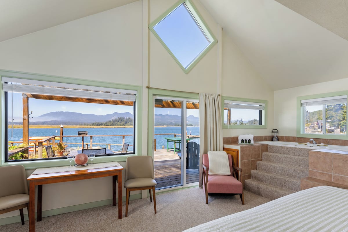 A room with a view of a lake, a dining area with a table and chairs, a sofa, a staircase, and a bathroom with a sink and mirror.
