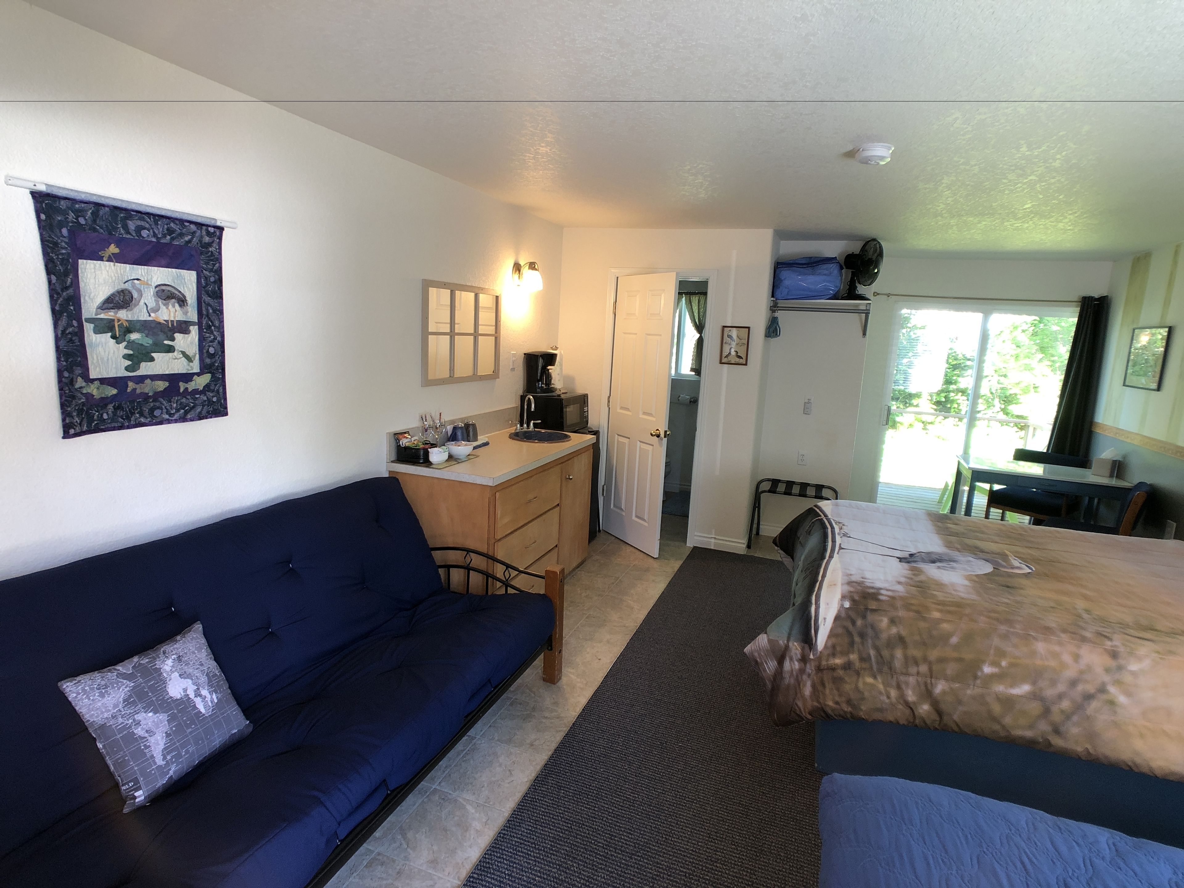 A compact room with a blue sofa, a small desk with a computer, a bed, and a view of the outdoors.