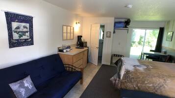 A compact room with a blue sofa, a small desk with a computer, a bed, and a view of the outdoors.