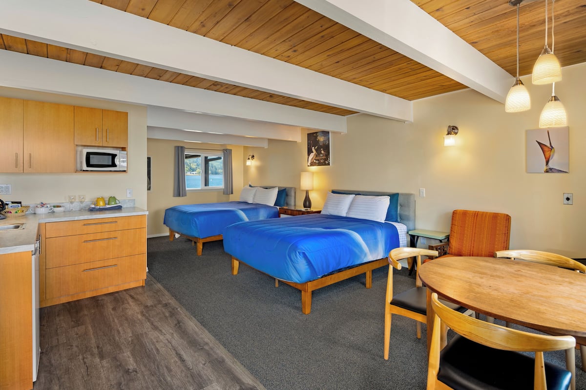 A room with two beds, a dining table, wooden cabinets, a microwave, and a ceiling with wooden beams.