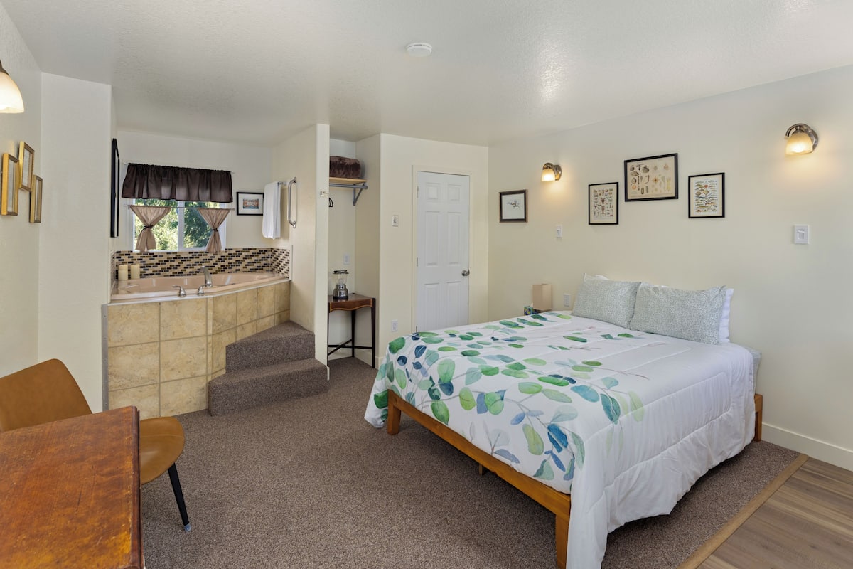 A bedroom with a bed, a desk, and a chair. There is a bathroom with a sink and a shower. The room has a carpeted floor and a door leading to another room.