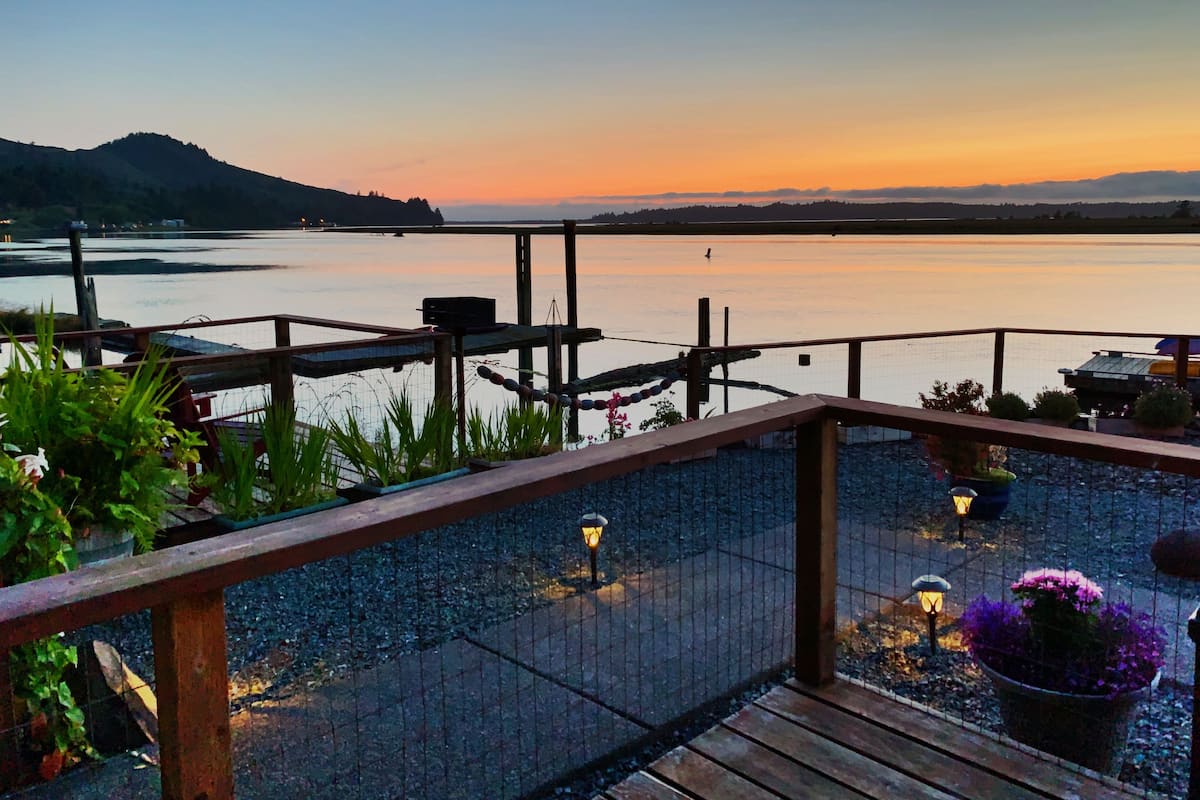 A waterfront deck with a railing, potted plants, and a view of the water and distant hills.