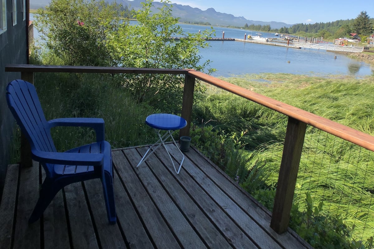 A wooden deck with a blue chair and table, overlooking a lake with boats and mountains in the distance.