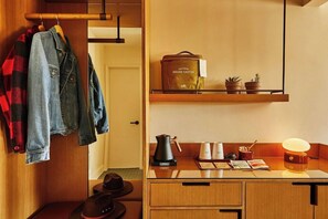 A hotel room with a wooden desk, a coffee maker, a shelf with a bag and plants, and hanging clothes.