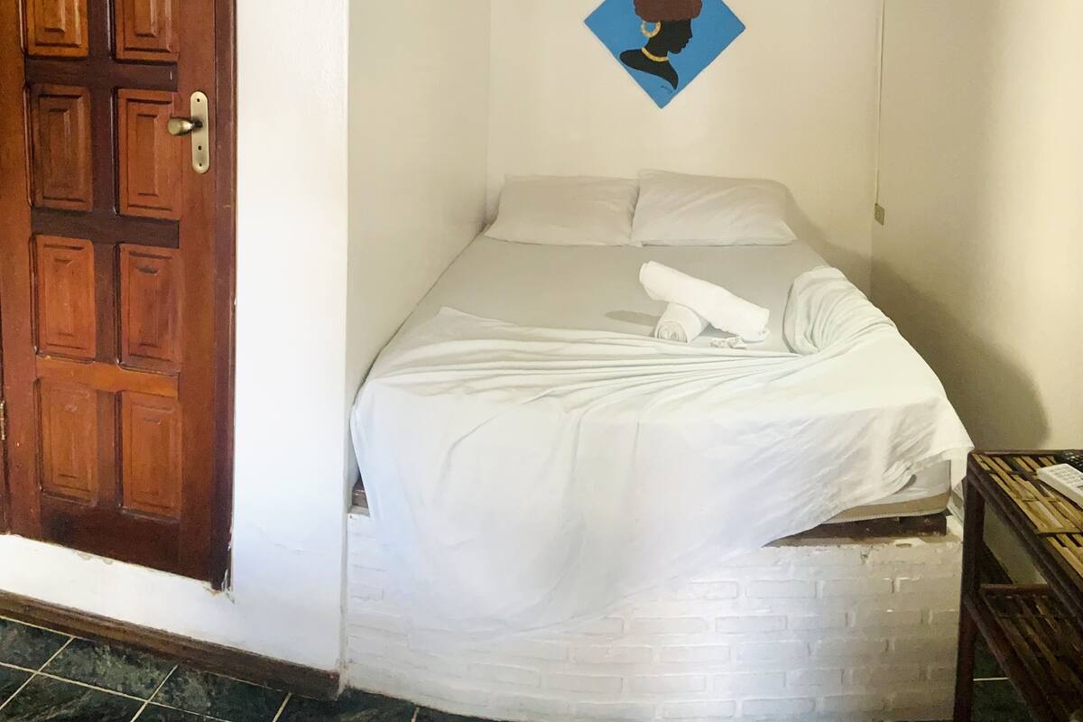 A room with a wooden door, a bed with white bedding, a blue wall decoration, and a brick-patterned wall.