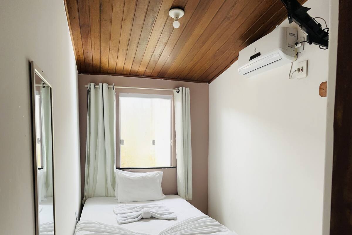 A small, neatly arranged bedroom with a single bed, a bedside table with a remote control and papers, a window with curtains, and an air conditioning unit on the wall.