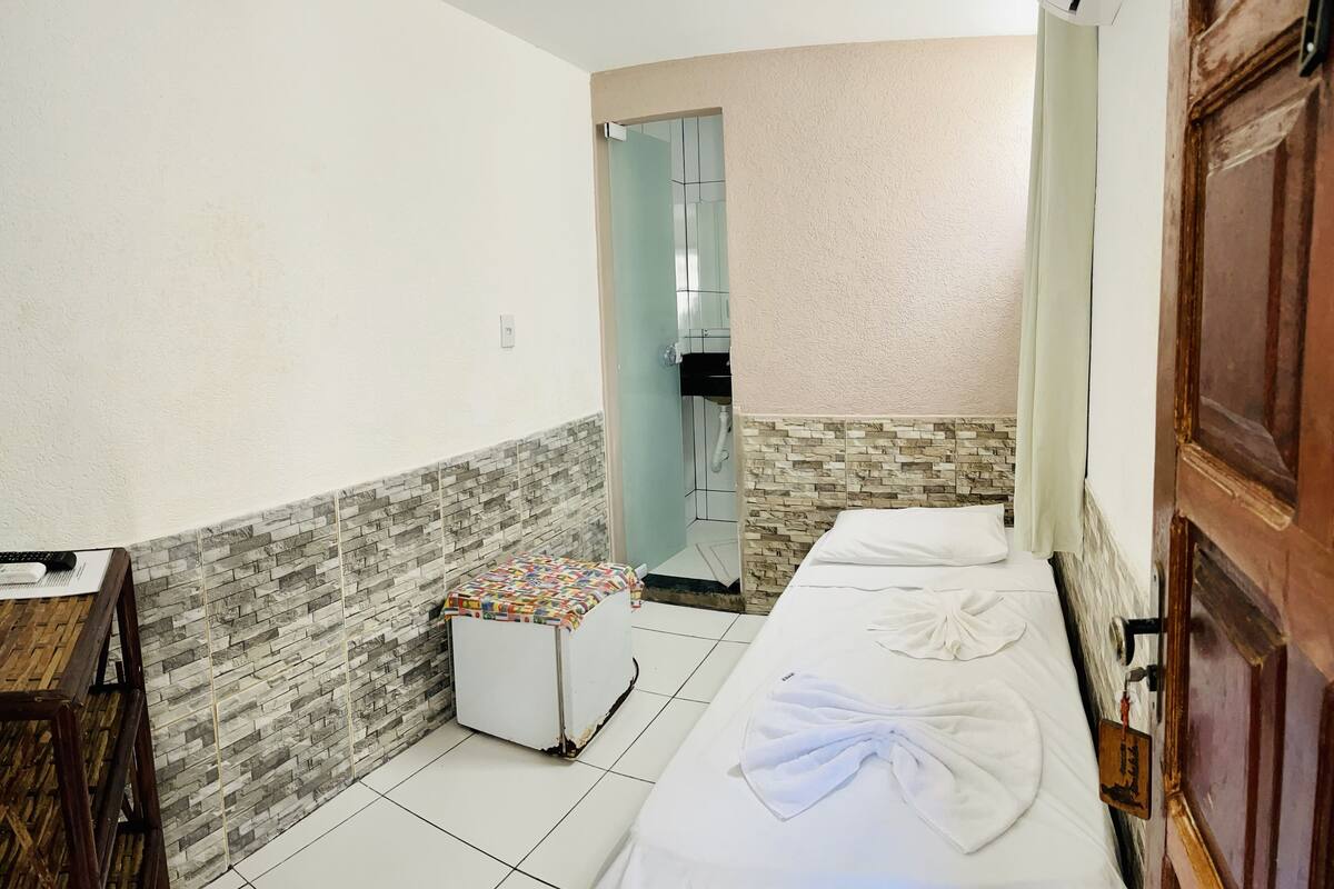 A small hotel room with a bed, a small table, and a bench. There is a bathroom visible through a glass door.