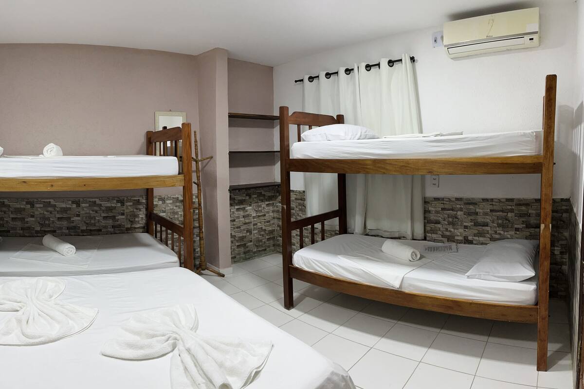 A room with three bunk beds, each with white bedding and towels neatly arranged. There is a wall-mounted air conditioner and a window with curtains.