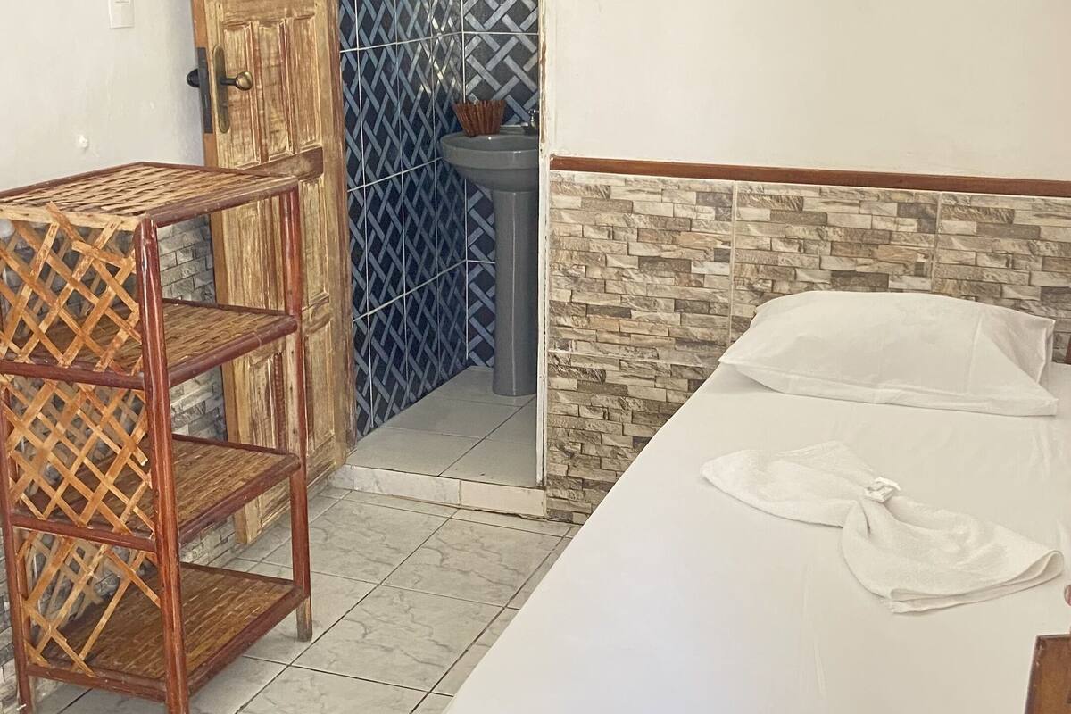 A room with a bed, a wooden chair, a bathroom with a sink, and a door leading to another room.