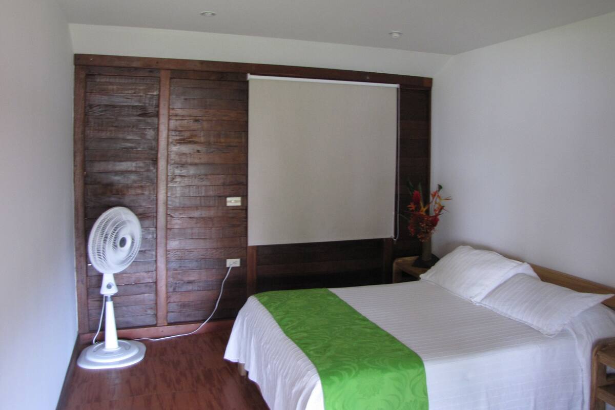 Une chambre avec une armoire en bois, un lit blanc avec une couvre-lit verte, une table de chevet blanche et un ventilateur blanc.