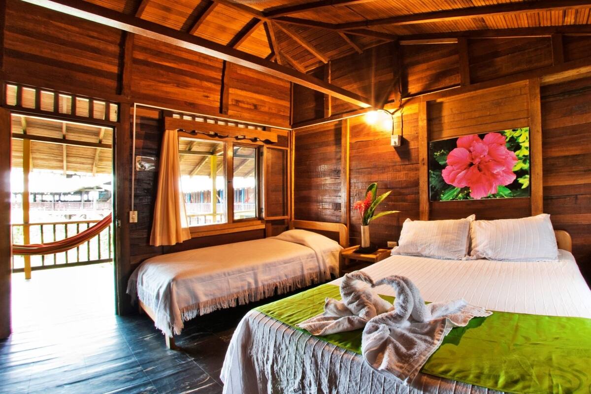 Une chambre dans un chalet en bois, avec deux lits, un hamac et une grande fenêtre donnant sur un paysage magnifique.