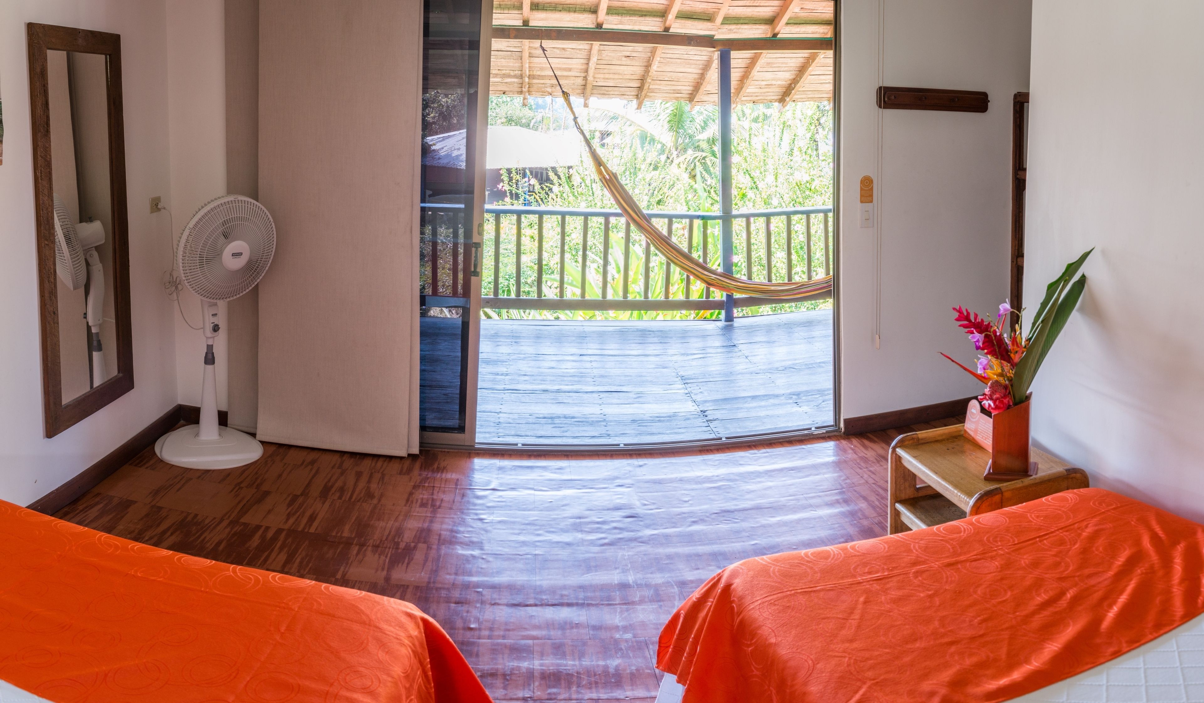 A room with two beds, a fan, a hammock, and a view of a wooden deck and greenery.