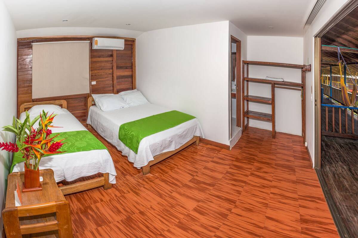 Une chambre de taille modeste, dotée de deux lits, d’un plancher en bois, d’une petite table ornée d’un vase de fleurs et offrant une vue sur un espace extérieur agrémenté d’une structure en bois.