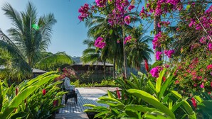 Una terraza de madera con una silla, rodeada de exuberante vegetación tropical y flores rosadas.
