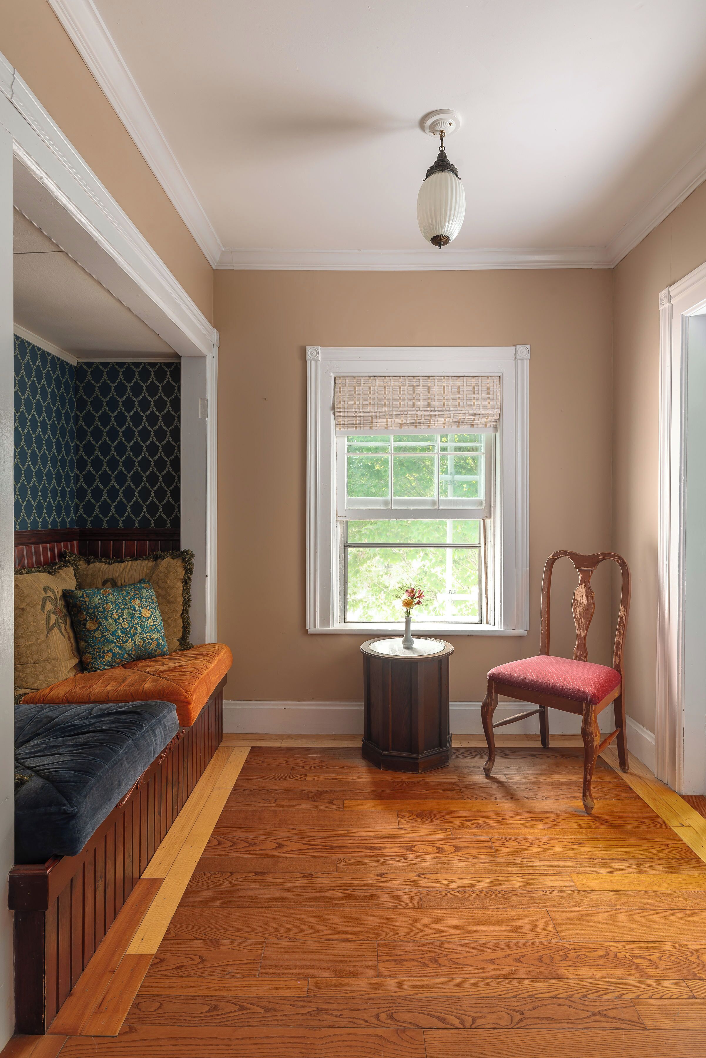 A room with a wooden bench, a chair, a small table with a vase, and a window with a white valance.
