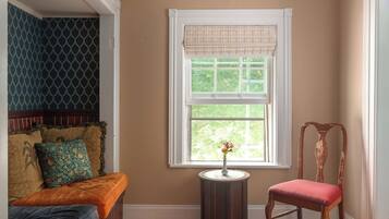 A room with a wooden bench, a chair, a small table with a vase, and a window with a white valance.