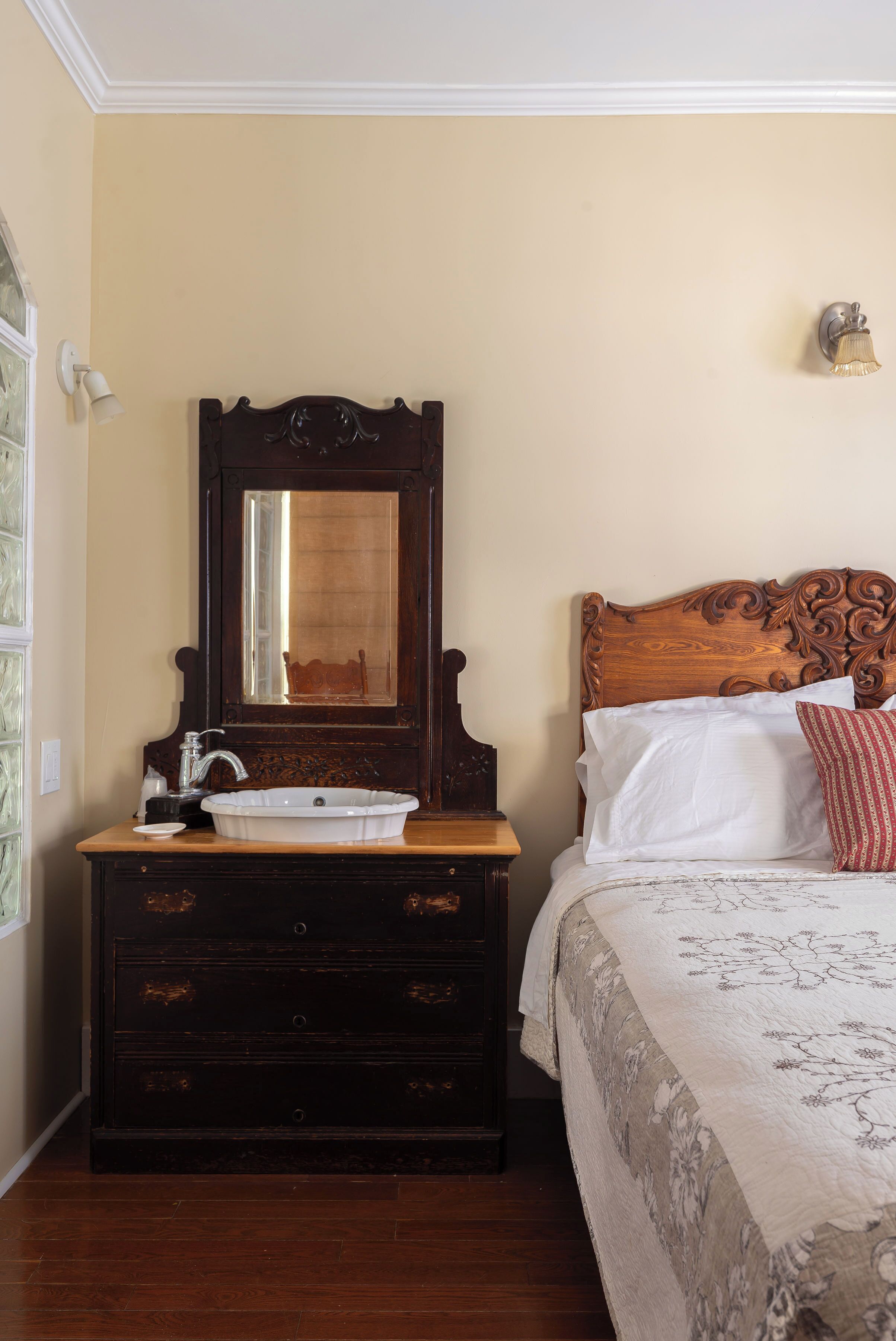 A room with a wooden dresser, a white sink, a mirror, a bed with white and red pillows, and a wall-mounted light fixture.