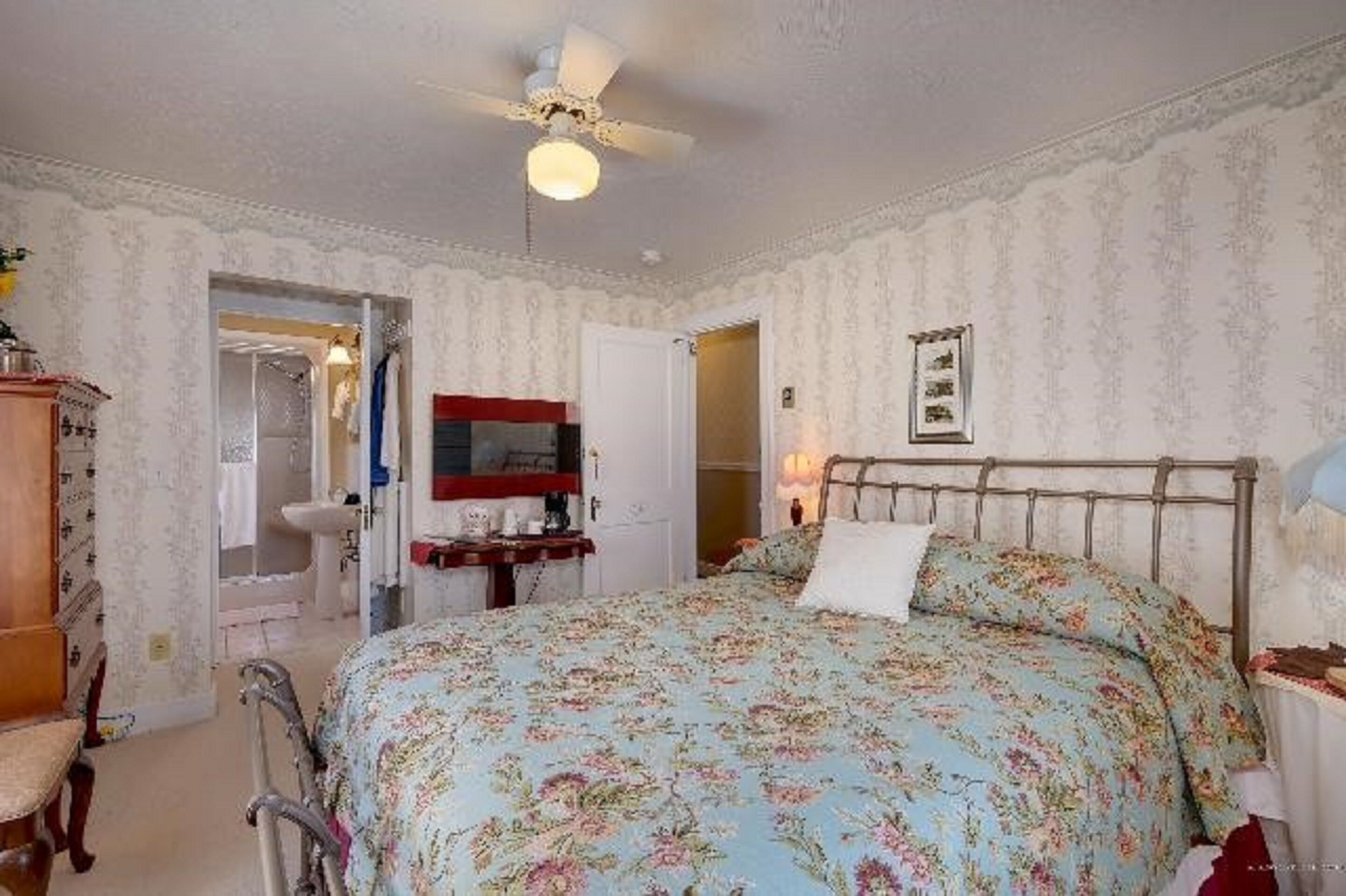 A bedroom with a floral bedspread, a ceiling fan, a wooden nightstand, and a mirror above the dresser.